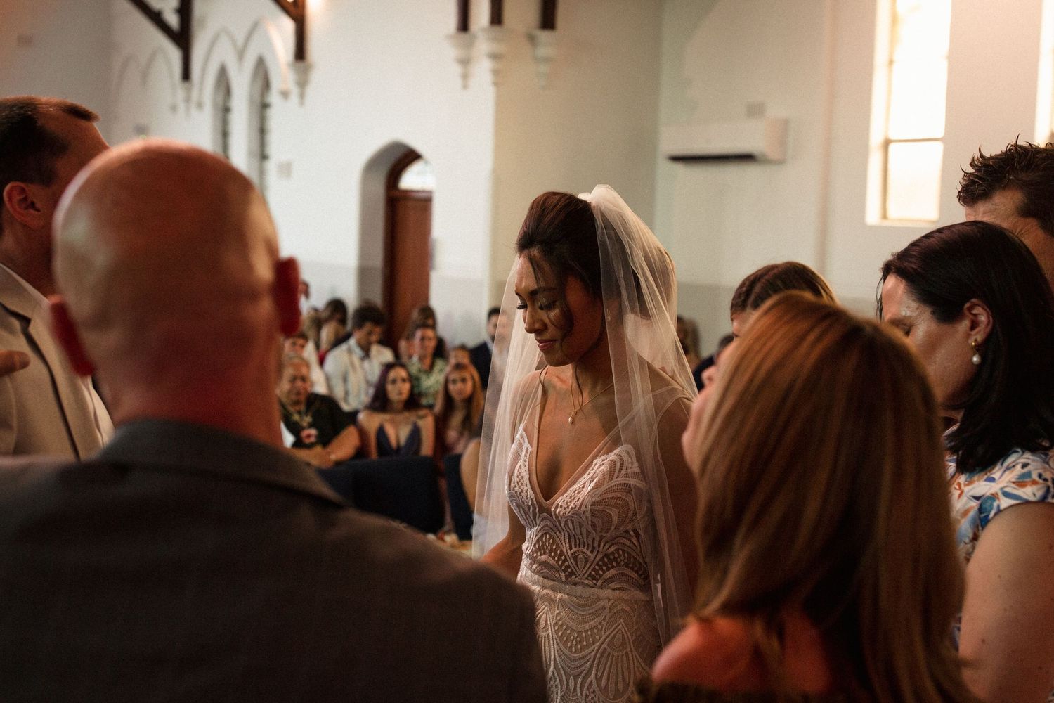 A bride in a detailed lace dress smiles during an intimate church ceremony moment.