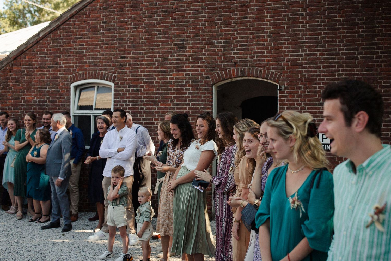 Wedding guests stand in line against a brick wall at an outdoor venue wearing an array of dresses and formal attire.