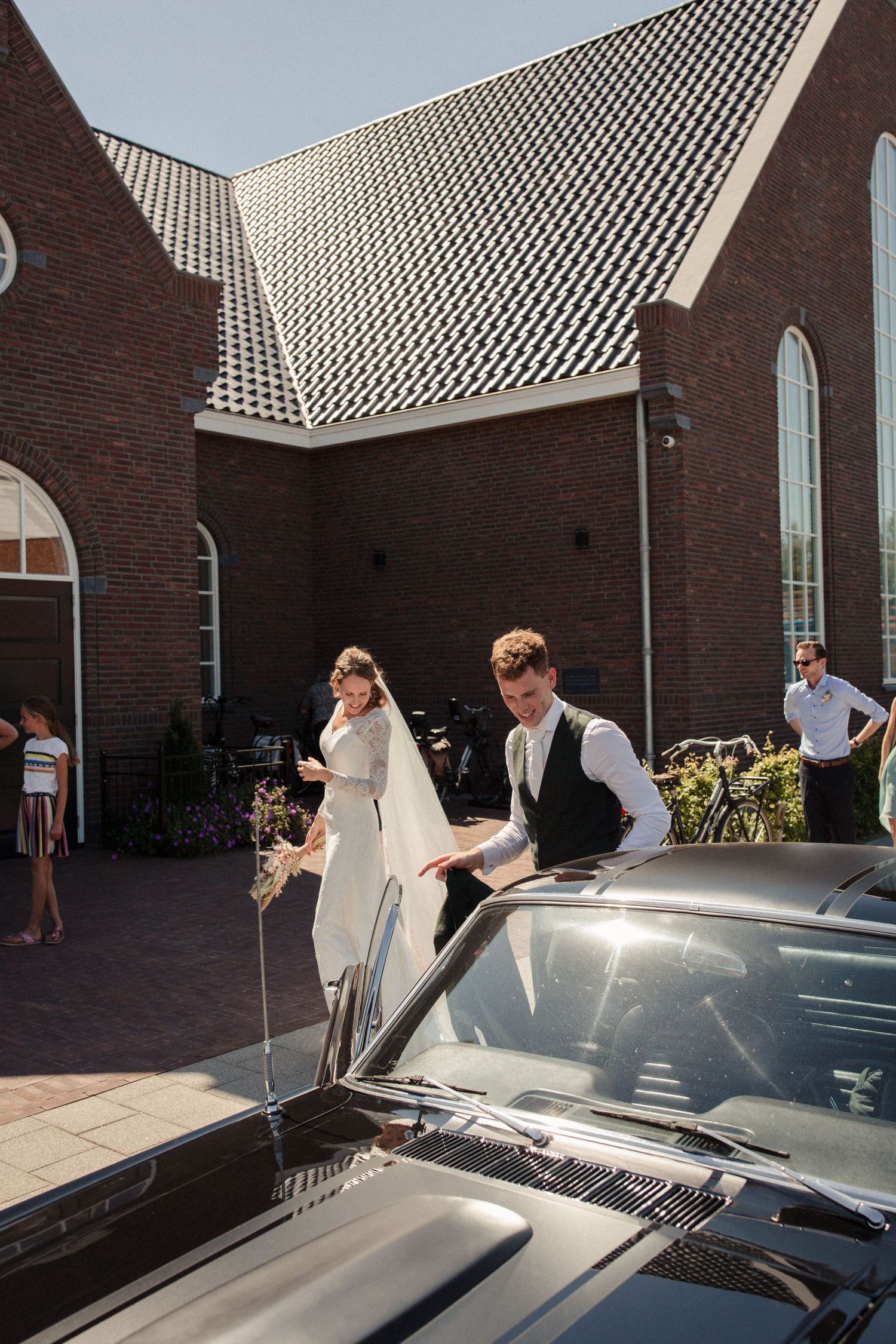 A couple in wedding attire next to a vintage car outside a brick church with distinctive patterned roof tiles.