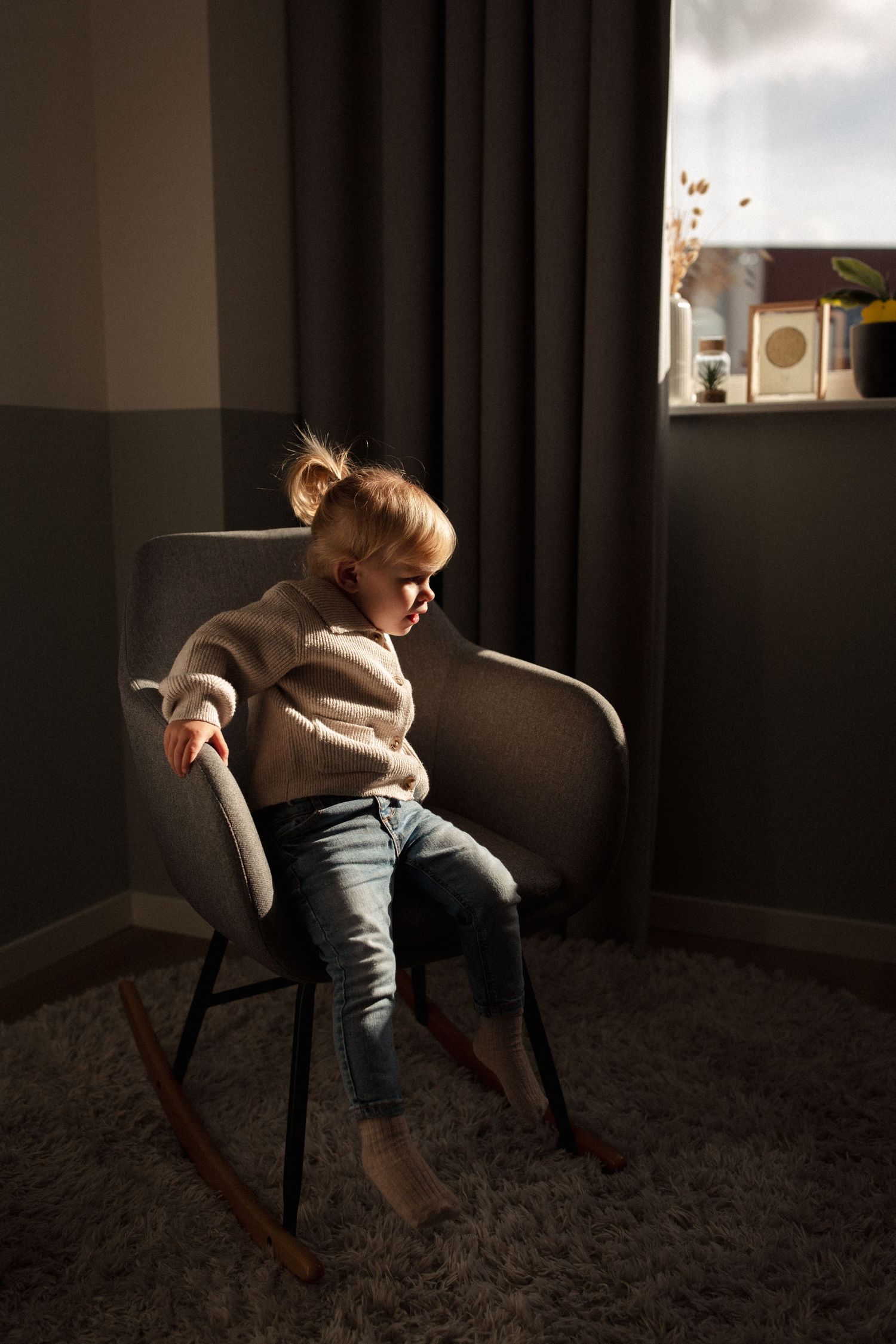 Small child in beige sweater sitting on grey rocking chair near window.