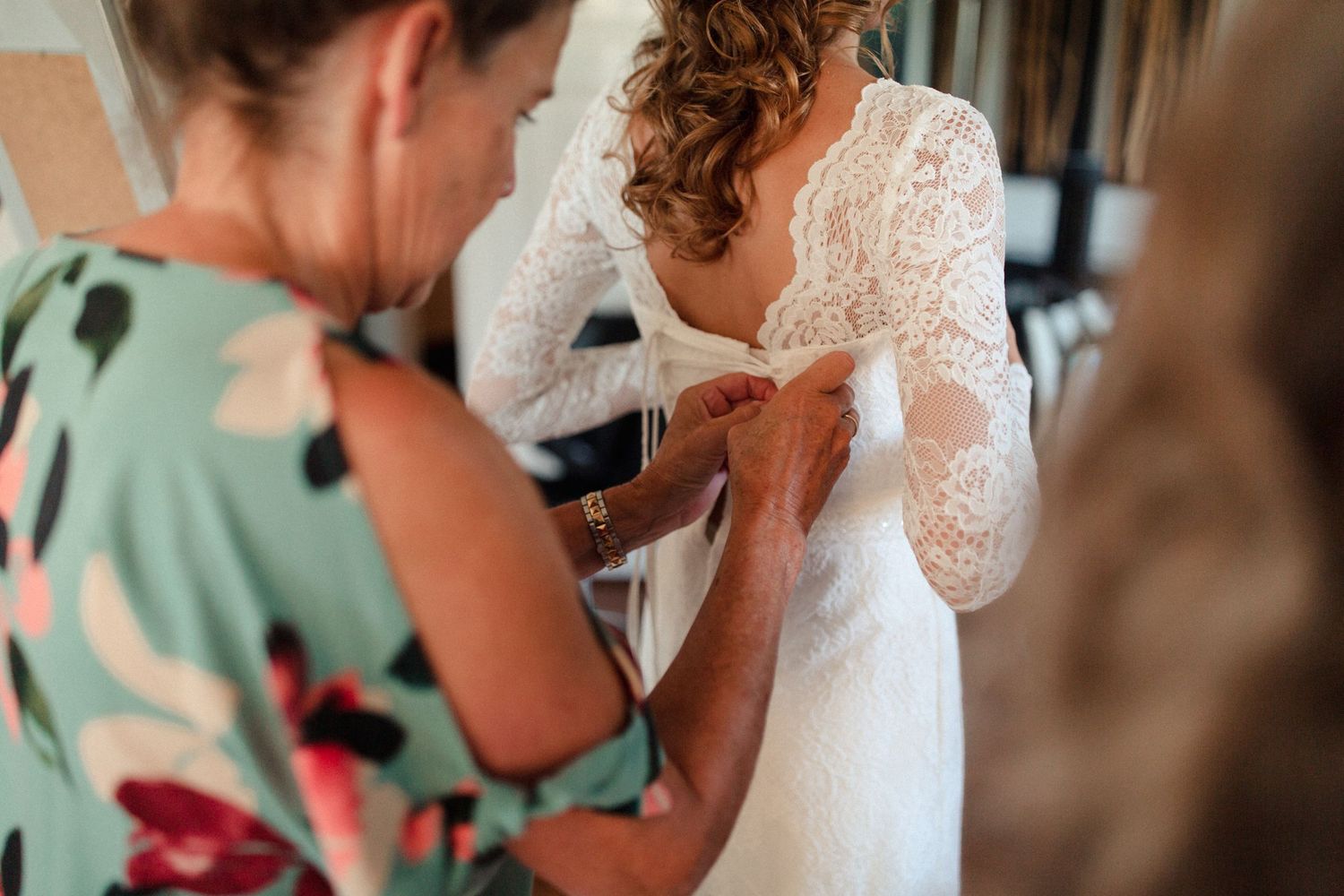 Woman in floral dress helps button up delicate white lace wedding gown during wedding preparation.