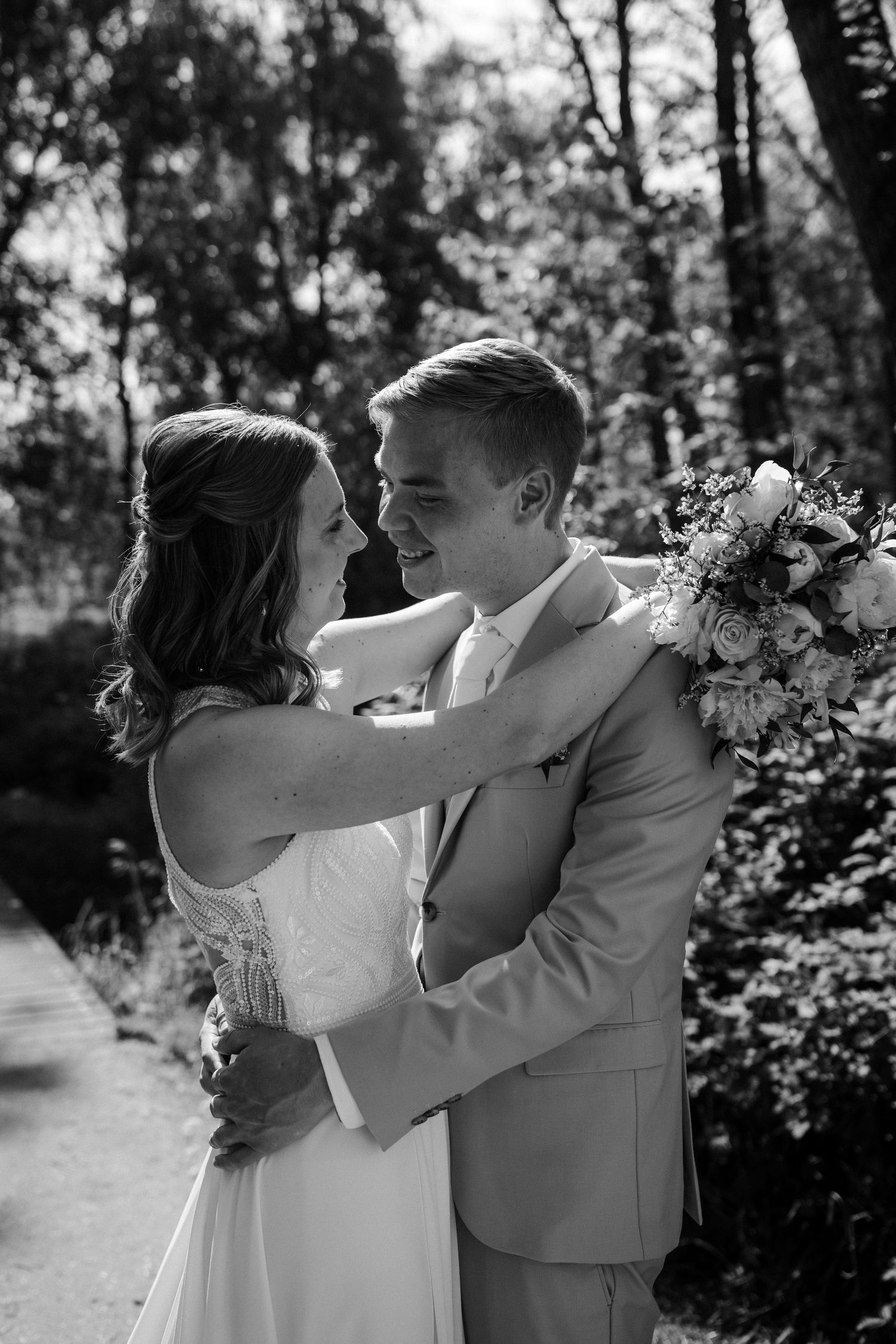A romantic black and white wedding photo of couple embracing outdoors with bride holding floral bouquet.