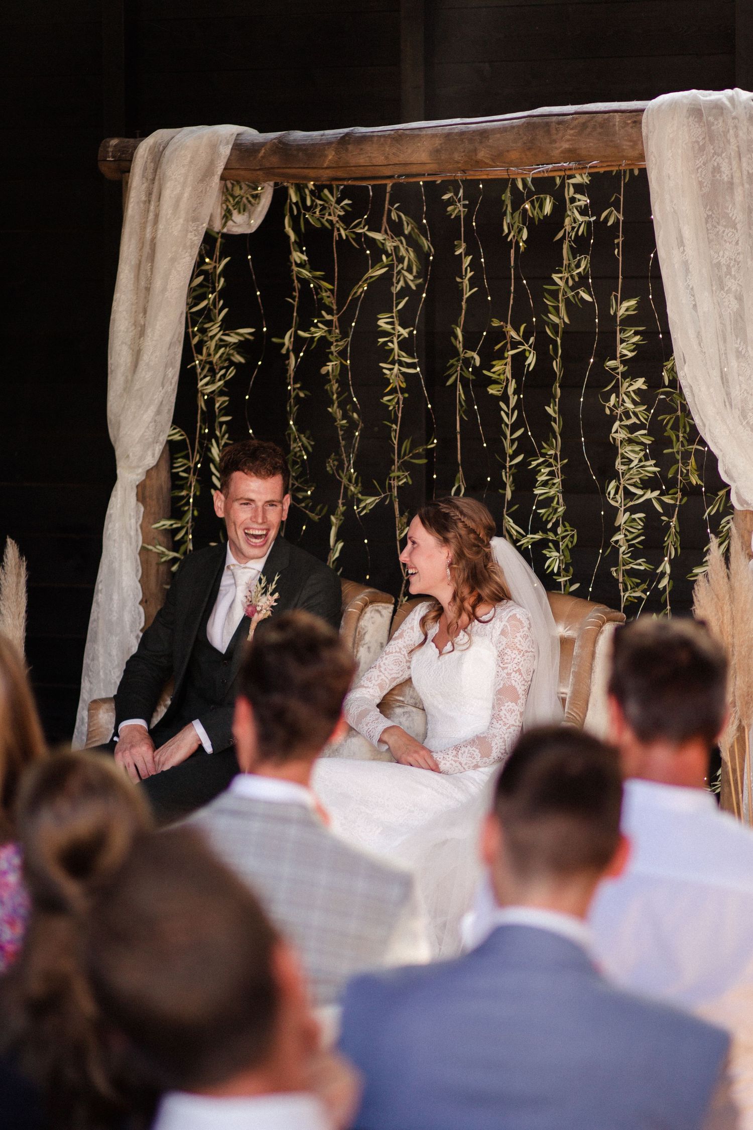 A rustic indoor wedding ceremony with draped fabric and hanging greenery garlands decorating the backdrop.