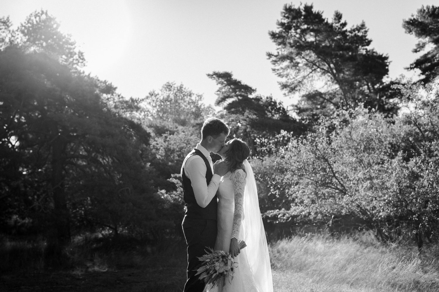 A romantic black and white wedding photograph showing a couple embracing outdoors with sunlight filtering through trees.