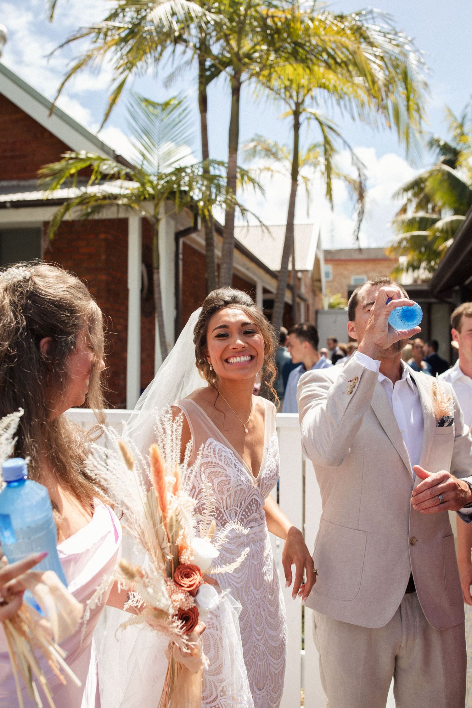 Wedding party celebrates outdoors with drinks under sunny palm trees.