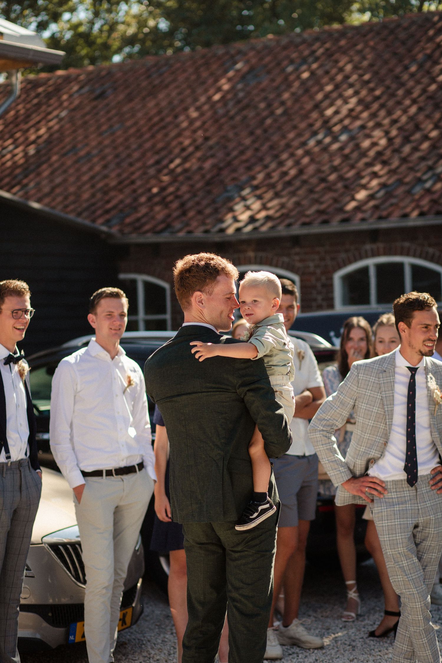 A group of wedding guests gathered outdoors near a building with a red tile roof on a sunny day.