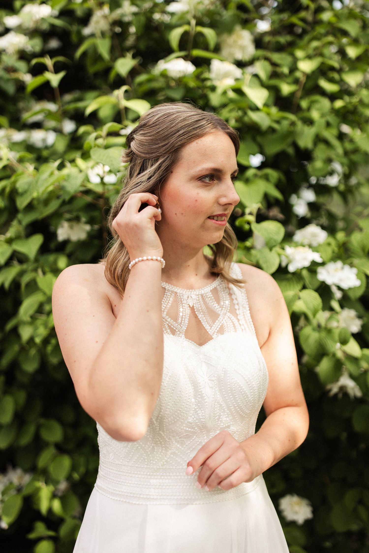 Person in a white lace wedding dress poses in front of flowering bushes while adjusting their earring.