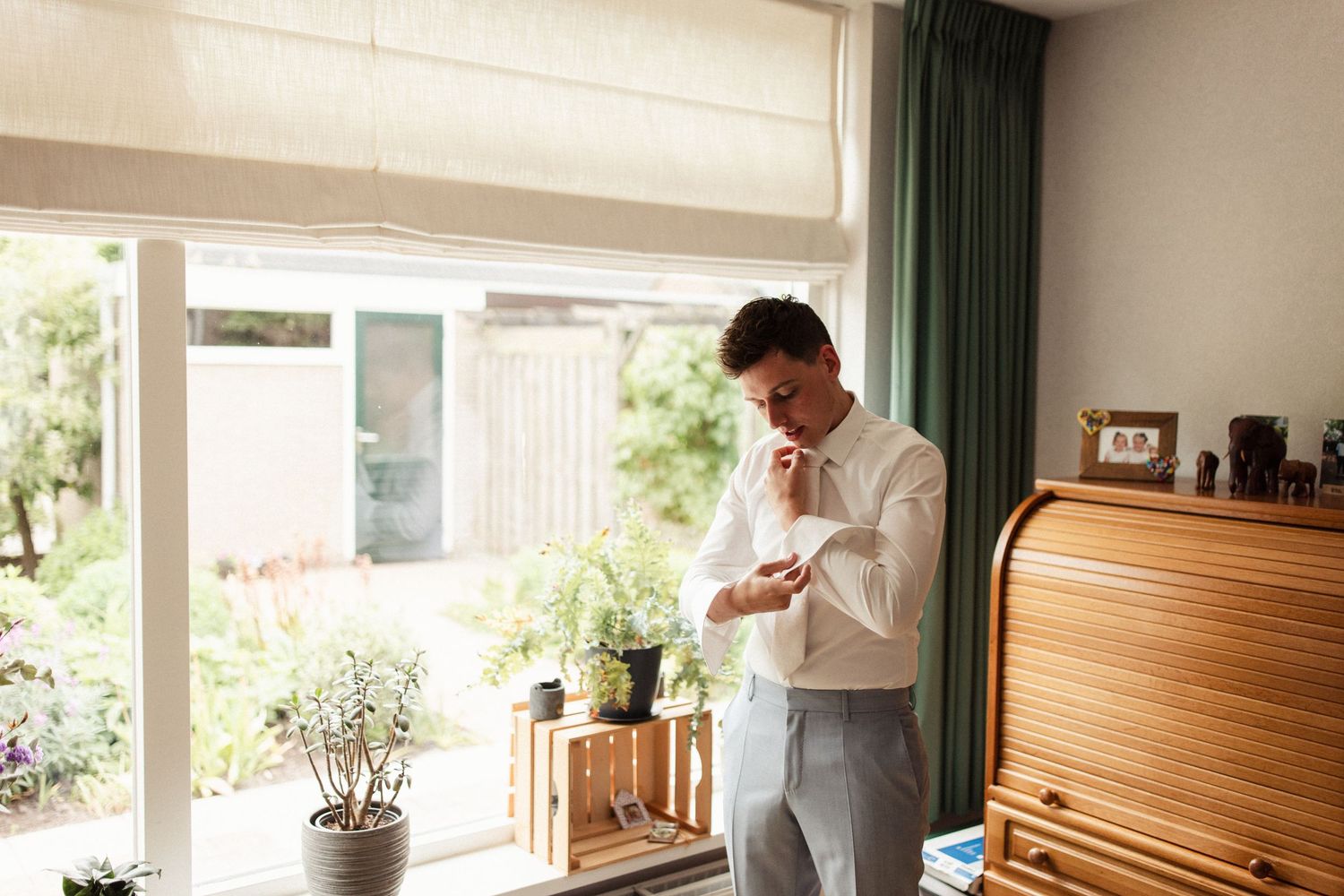Person in white shirt stands by window getting ready in morning light with plants and curtains.