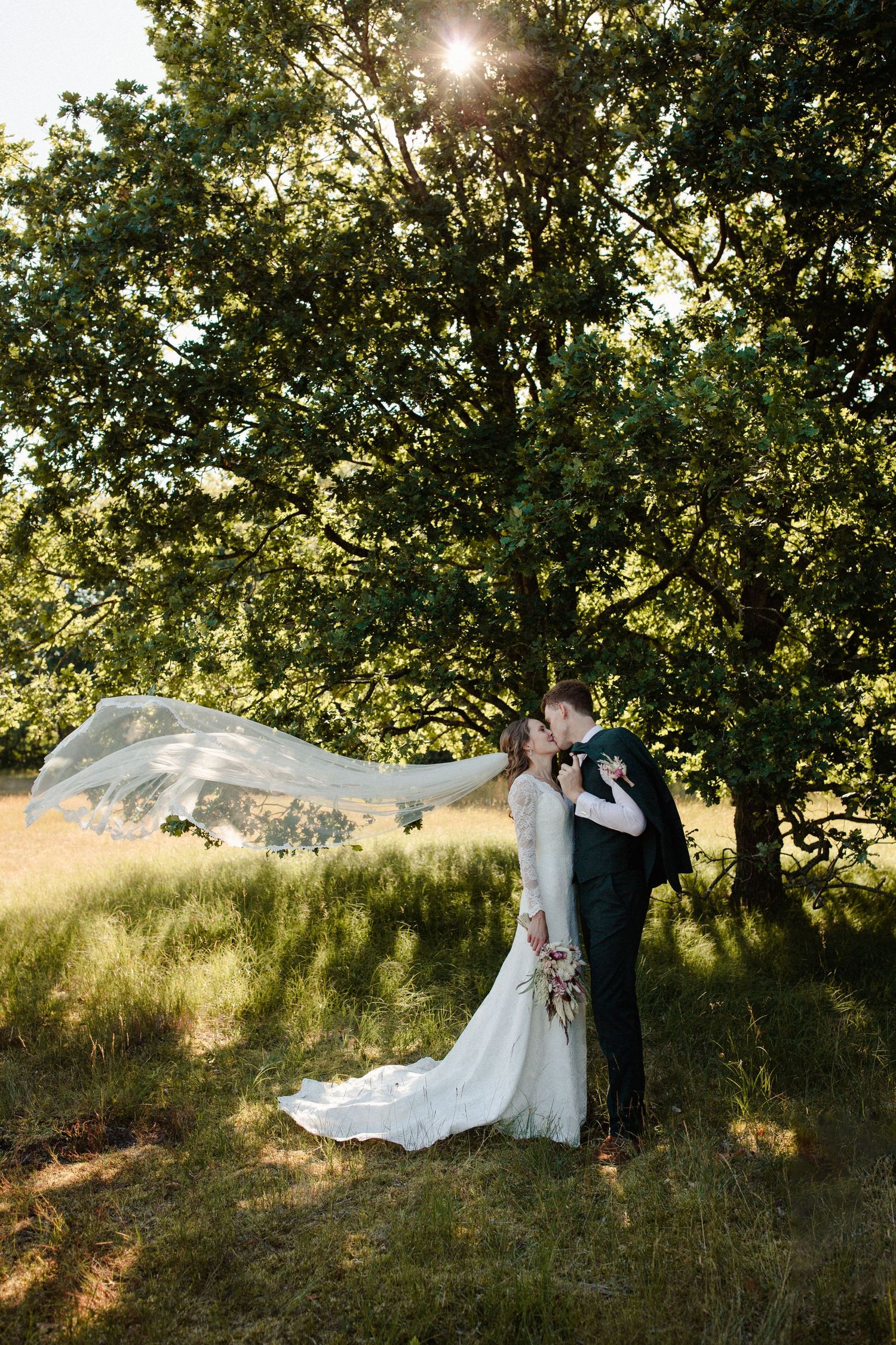 A couple embraces outdoors in a scenic meadow with sunlight filtering through trees while a white veil flows in the breeze.