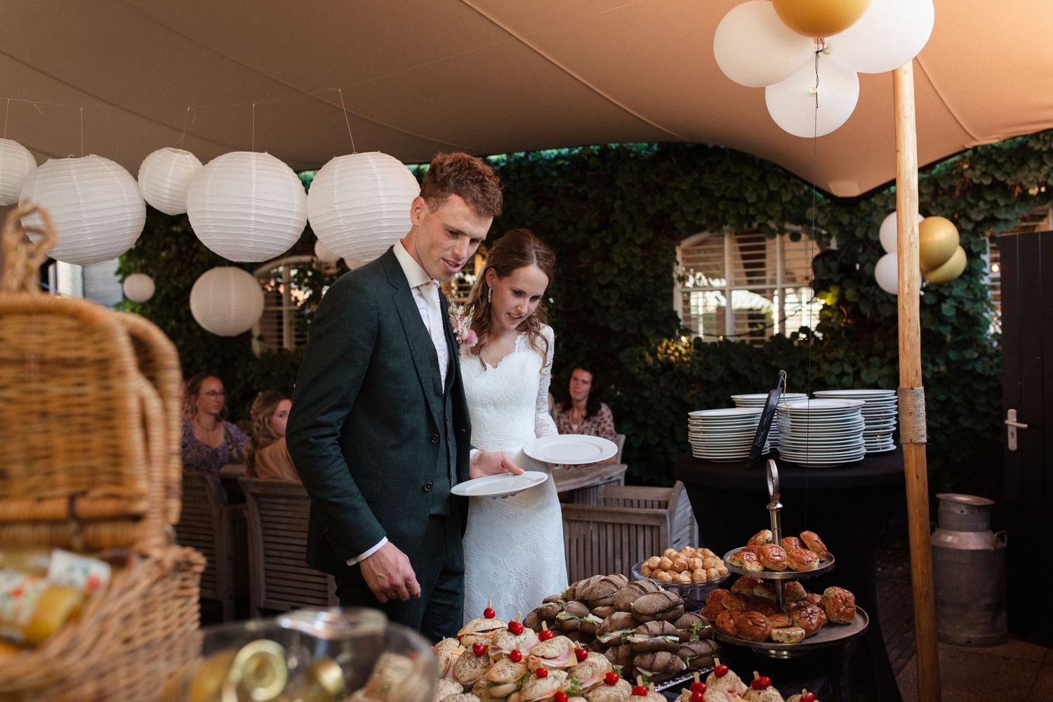 A couple in formalwear looks over a buffet table decorated with white balloons at an outdoor reception venue.