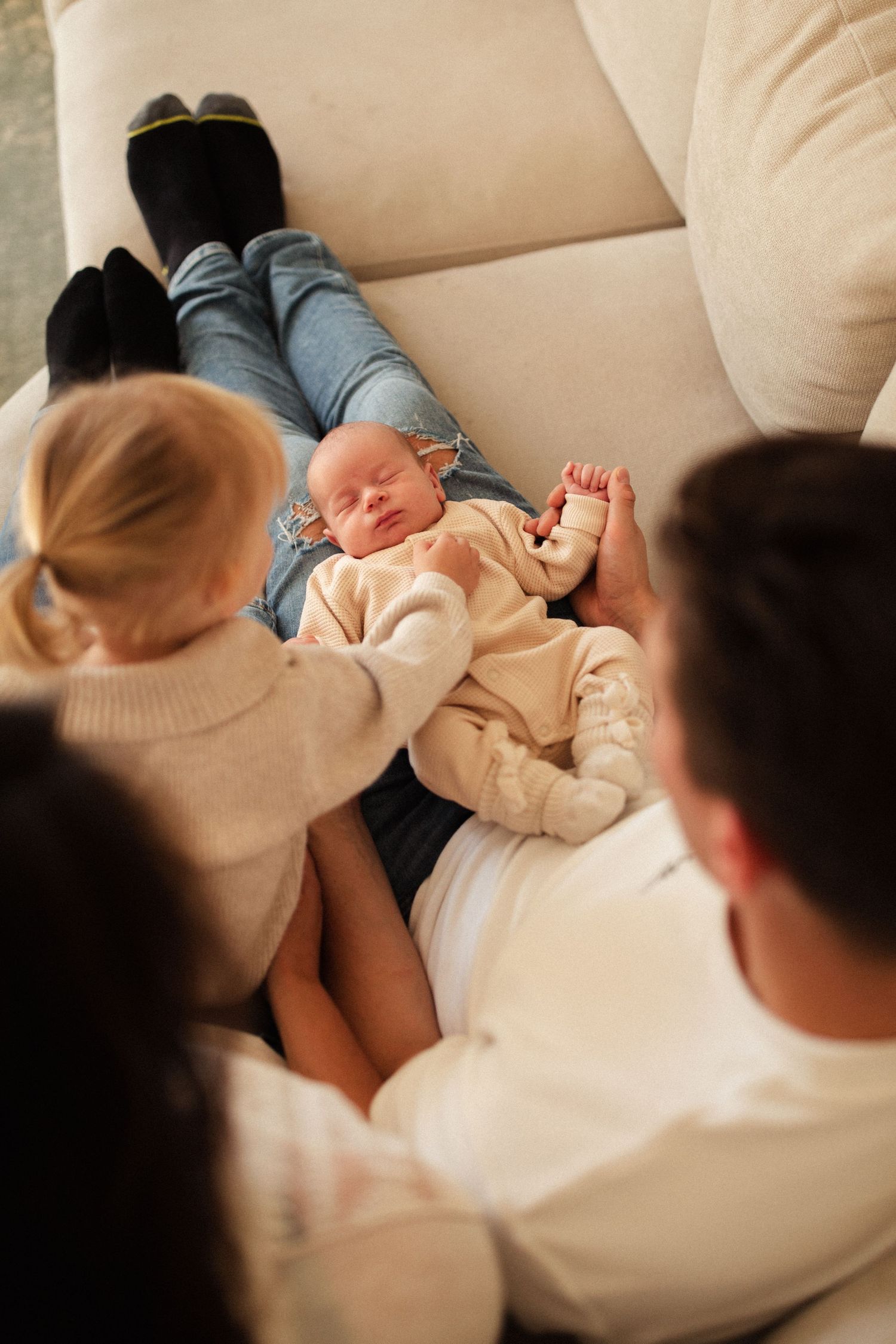 Family lounging together on cream colored sofa with sleeping infant in center.