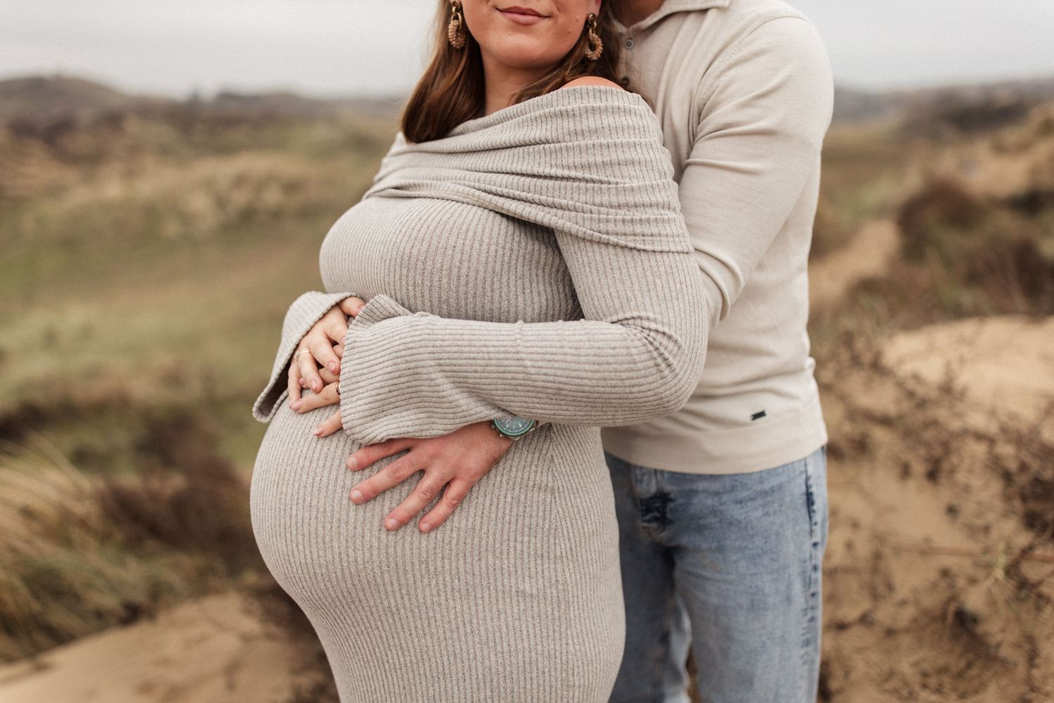 Man omarmd vrouw met zwangere buik. Ze staan ergens tussen de duinen.