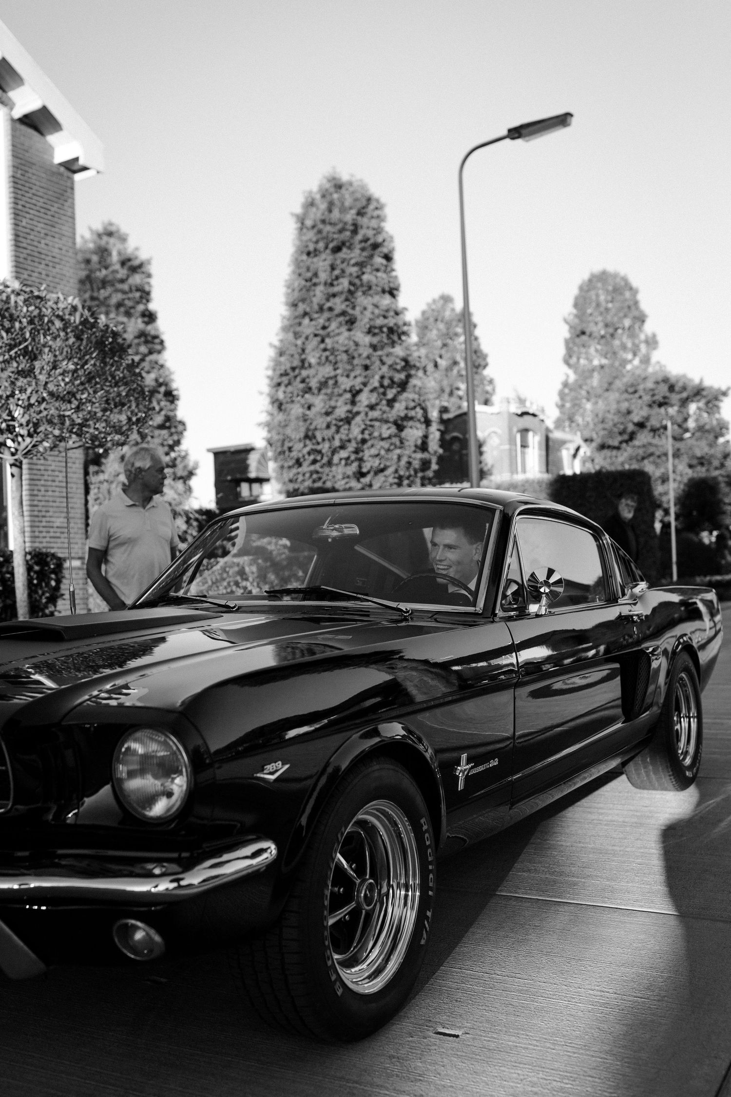 Classic black Ford Mustang fastback parked on street with tall trees in background in black and white photography.