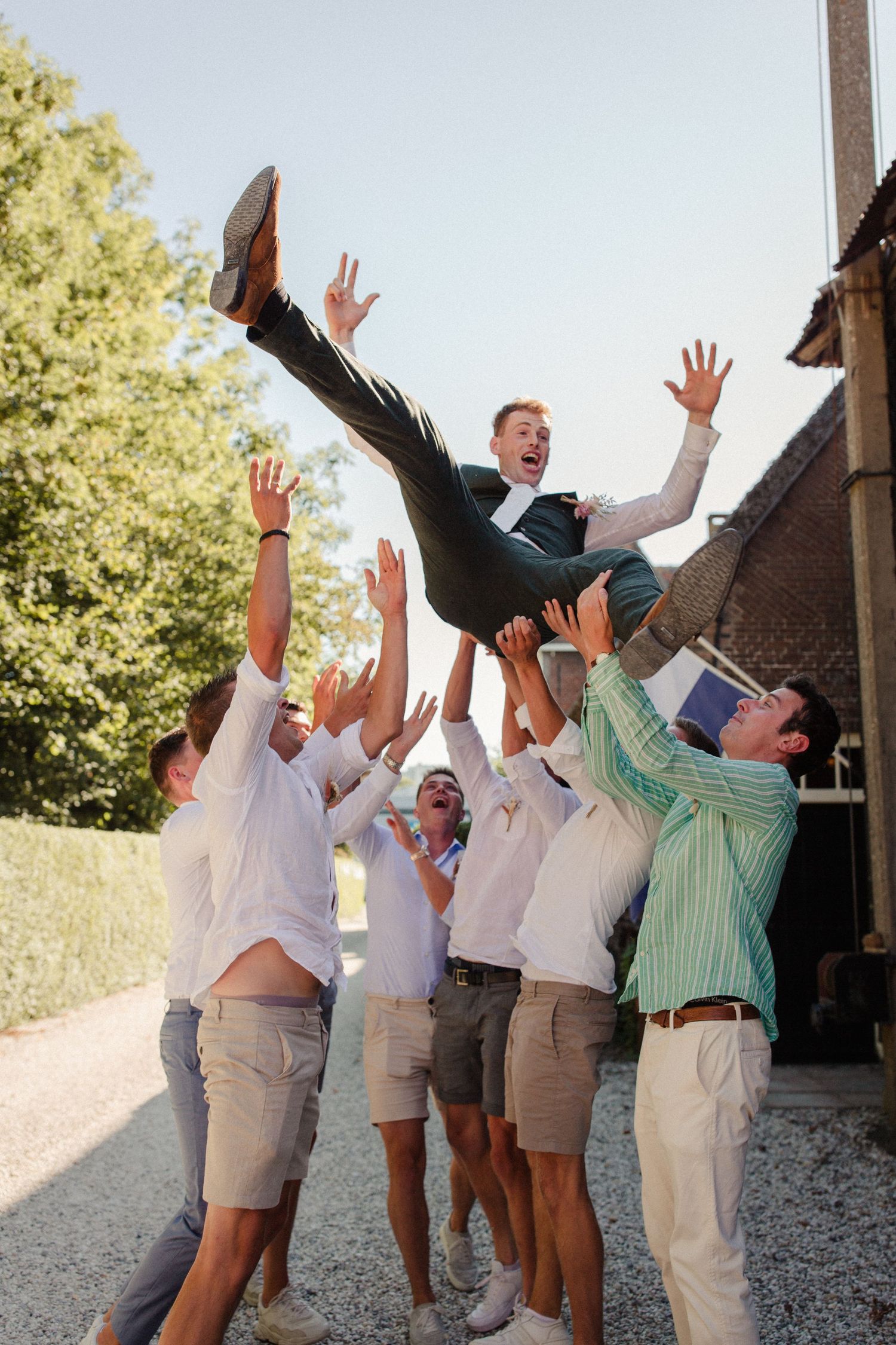 Groomsmen in khaki shorts and light colored shirts toss someone up in the air during outdoor wedding celebration.