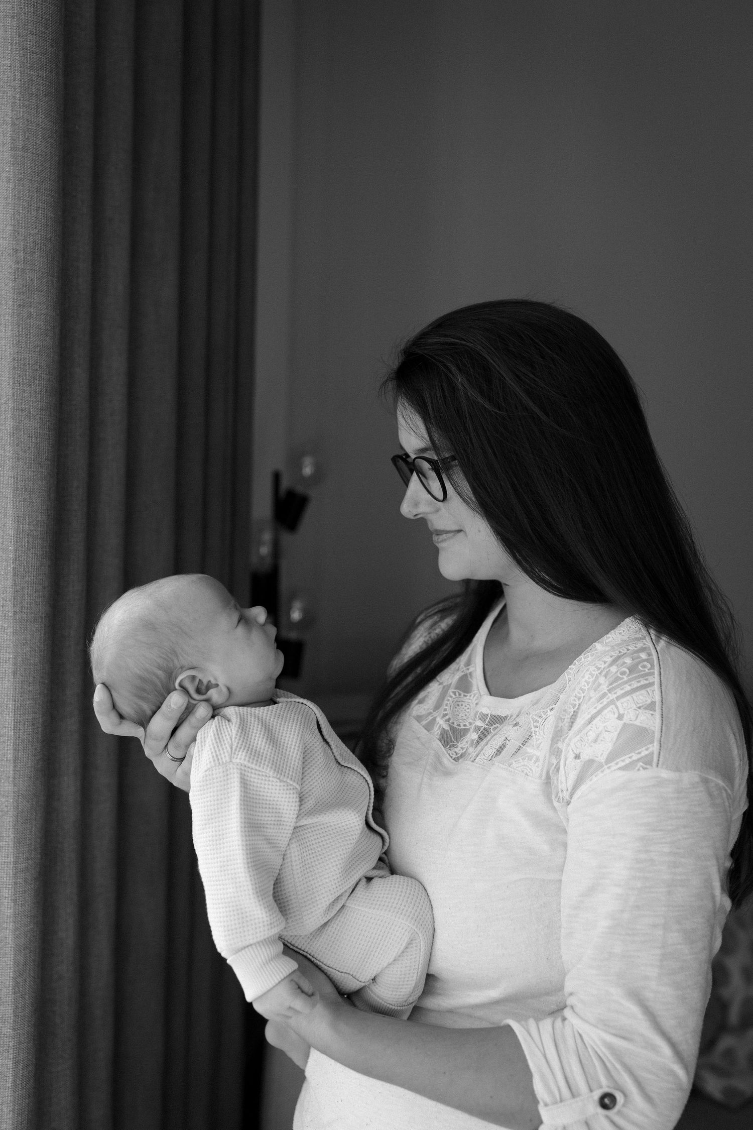 Woman in white lace holding infant in neutral clothing by window.