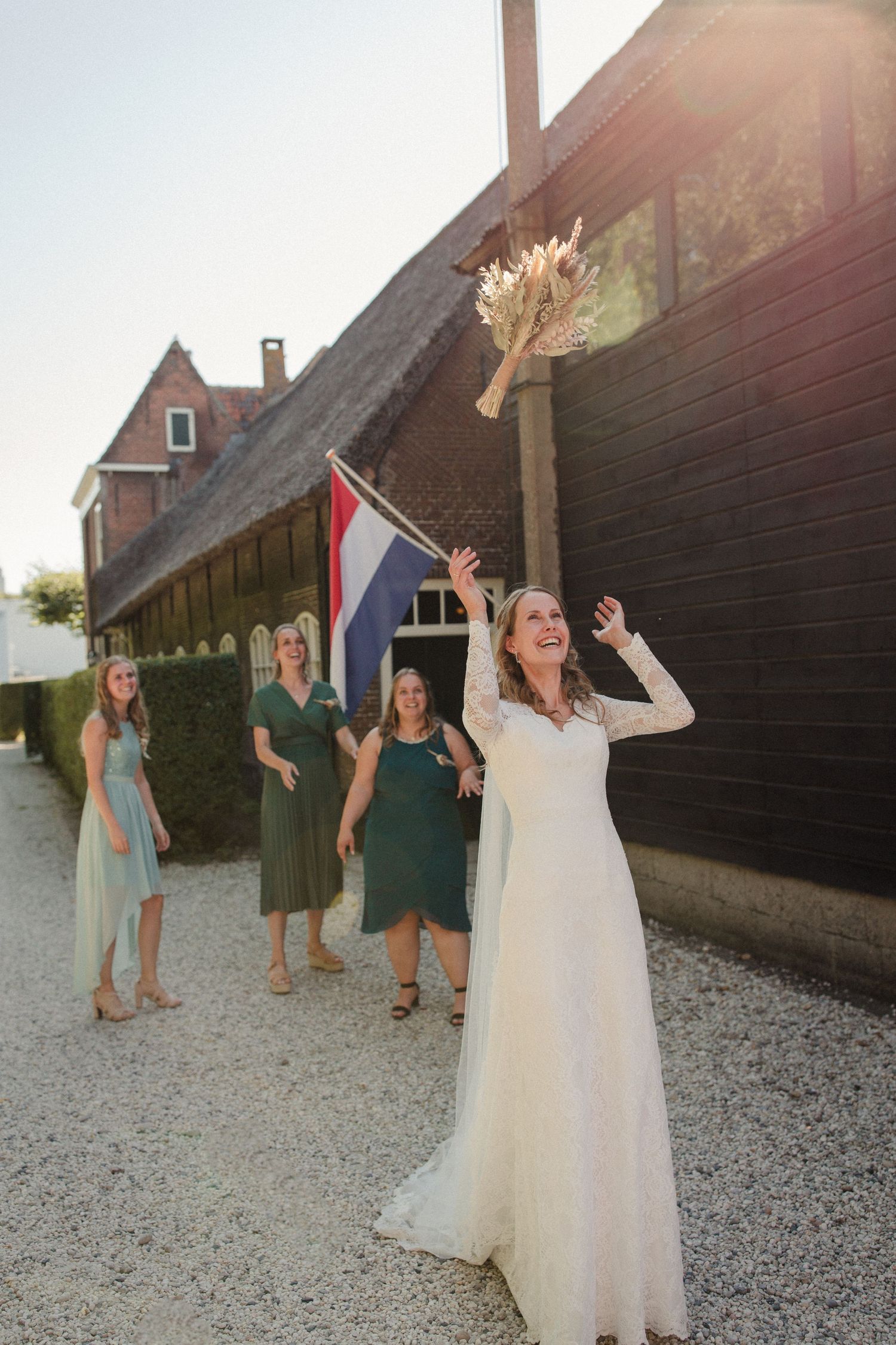 Wedding party celebrating outdoors by a rustic building with Dutch flag and sunlit backdrop.