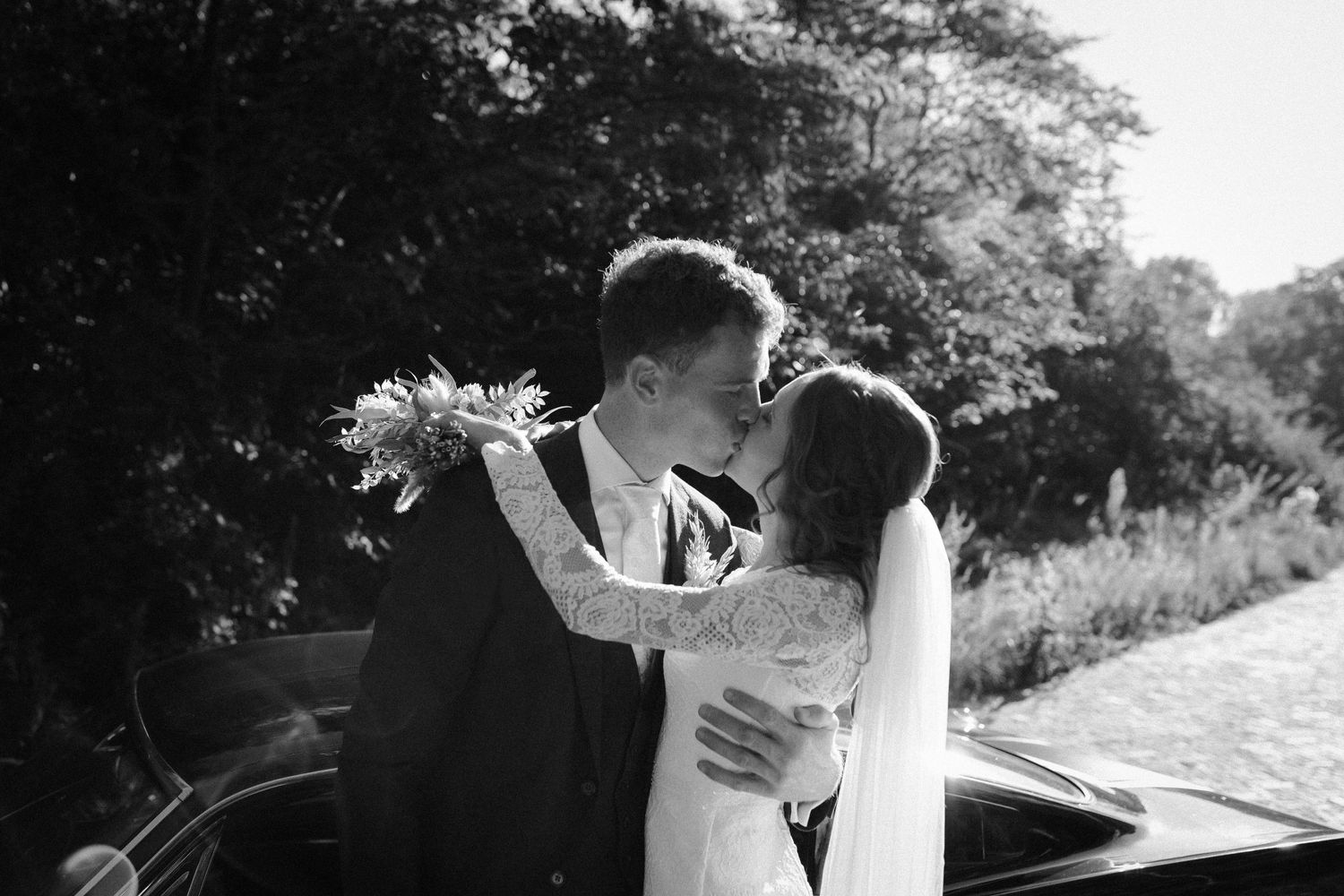 A romantic black and white wedding photo of a couple embracing and kissing alongside a vintage car.
