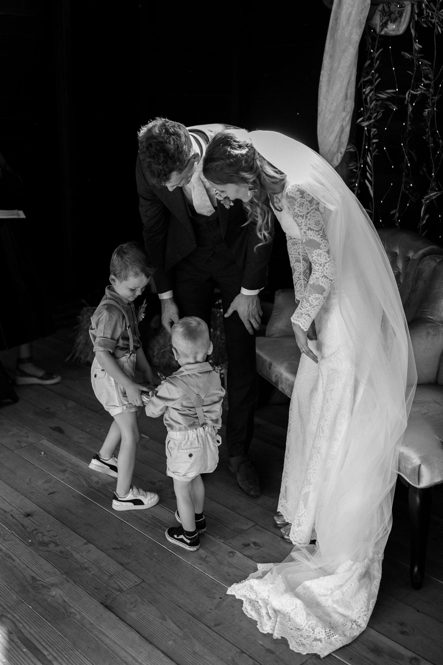 A bride in a flowing lace gown bends down to interact with two small children at a wedding celebration.