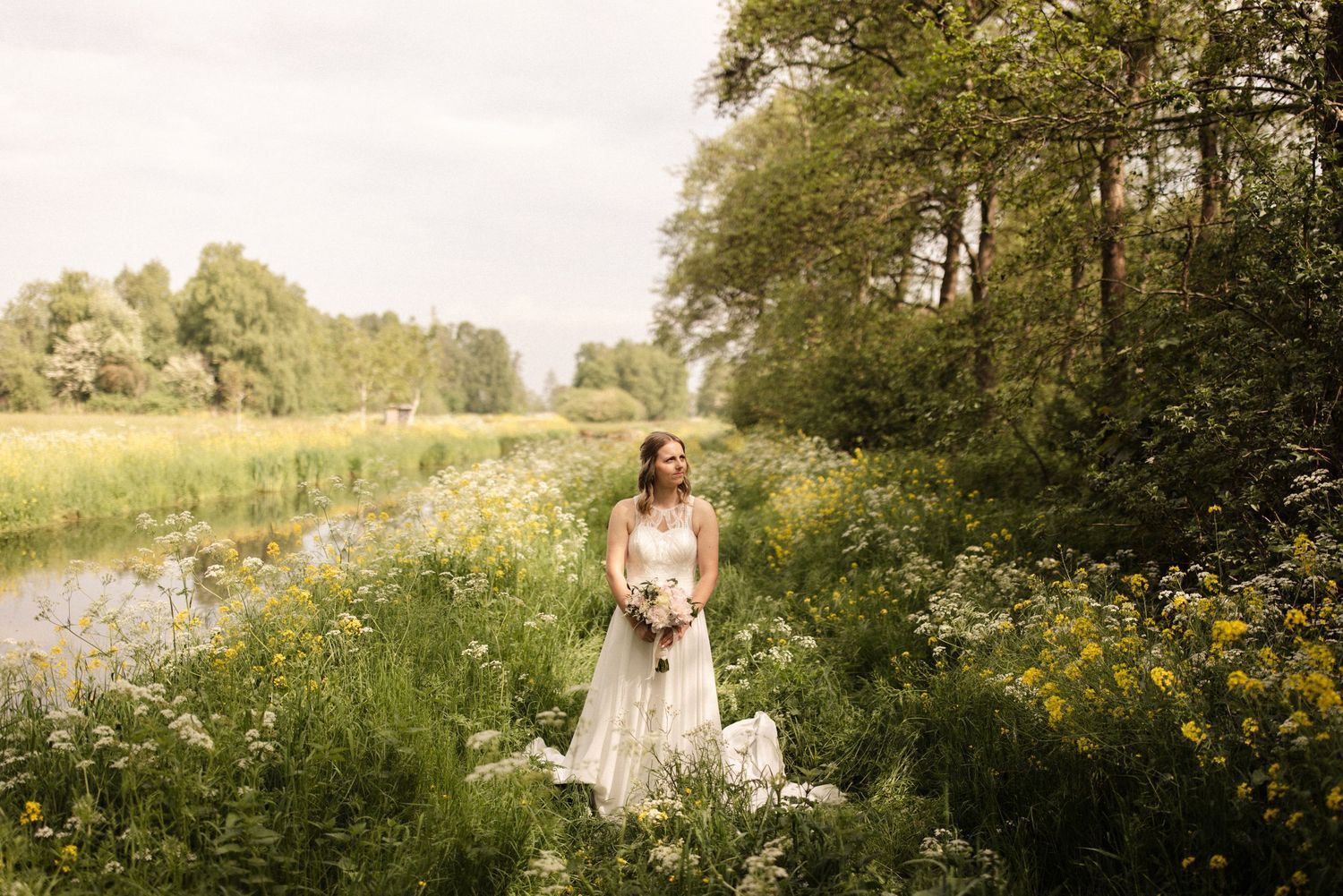 A bride in a white dress holds a bouquet while standing in a sunlit meadow with wildflowers and trees.