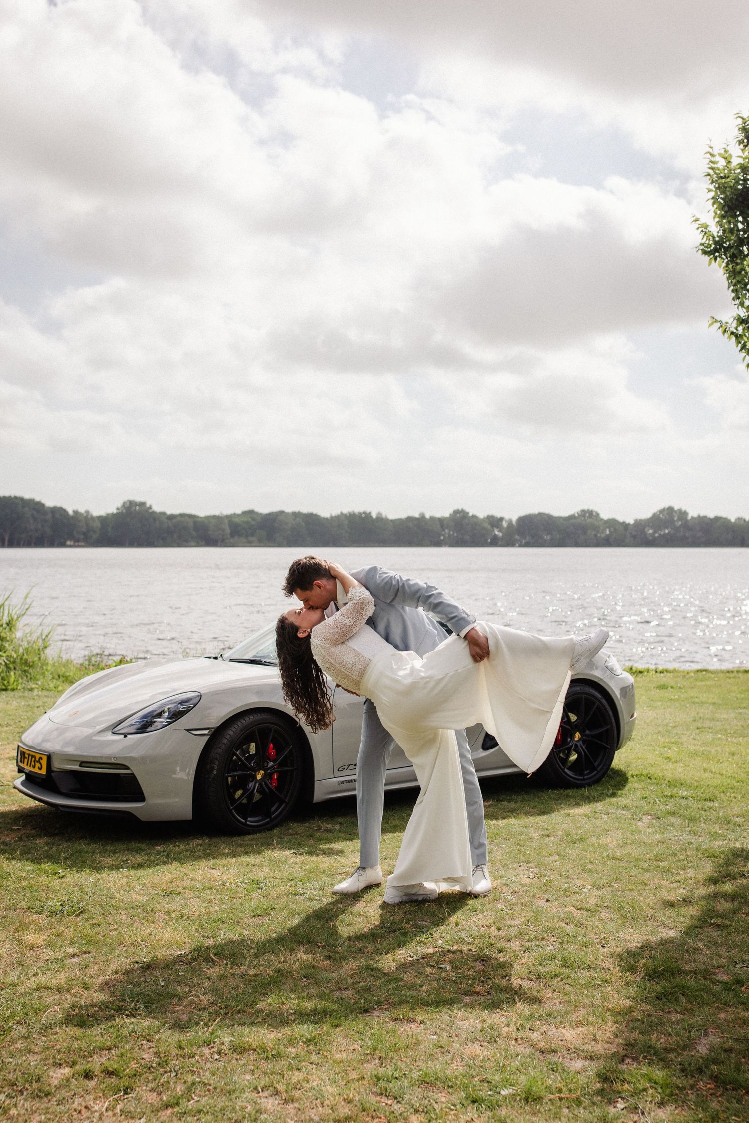 Bruidspaar die een dramatische kus doet voor hun trouwauto (porsche), met de Zegerplas als achtergrond