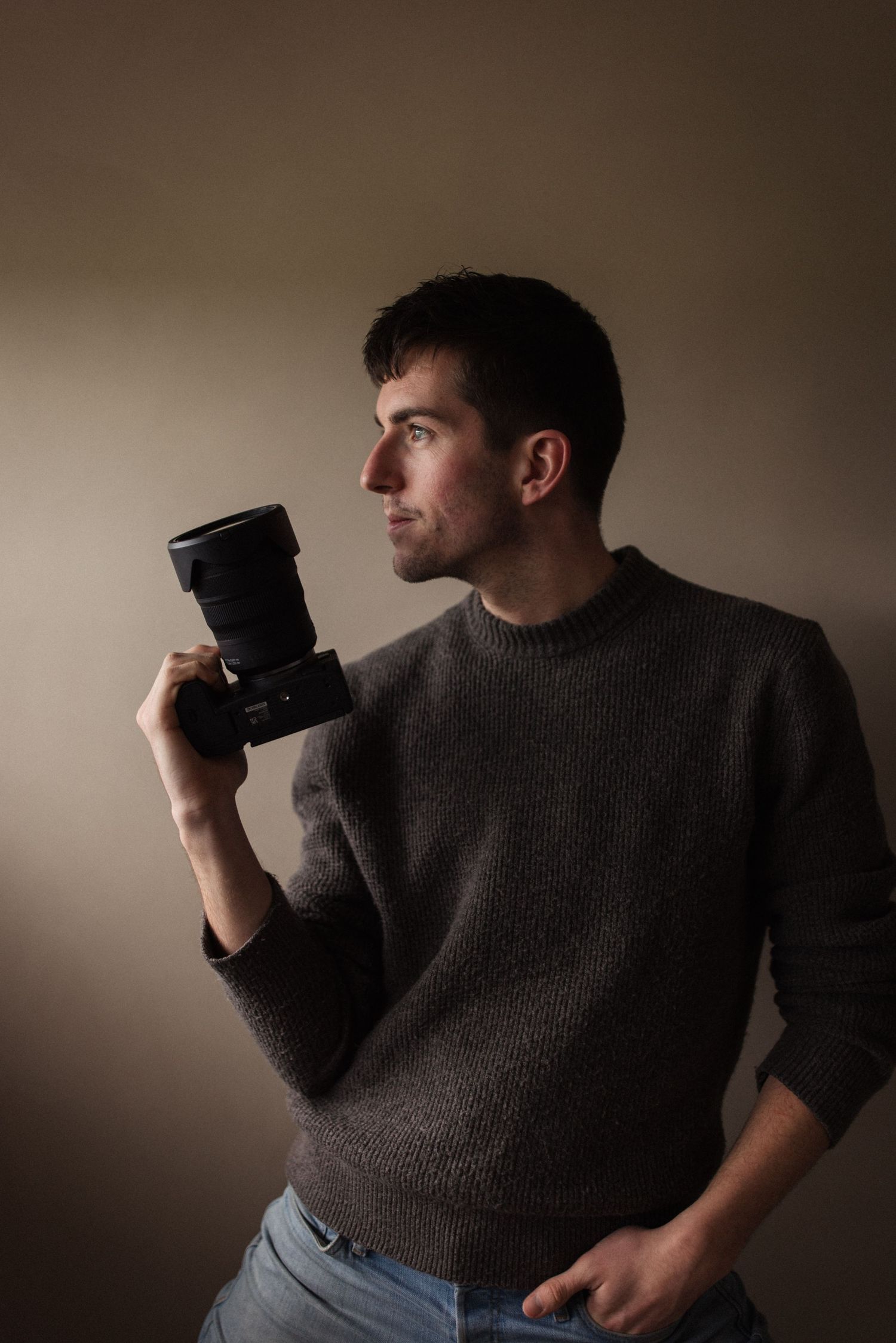 A photographer in a brown sweater holds a professional camera while looking to the side in moody lighting.