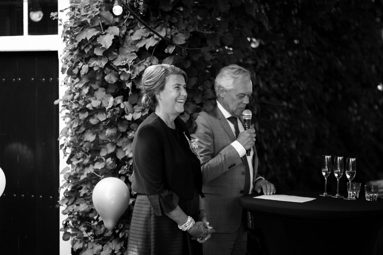 Two people stand at a podium with drinks and ivy backdrop at a formal black and white event.