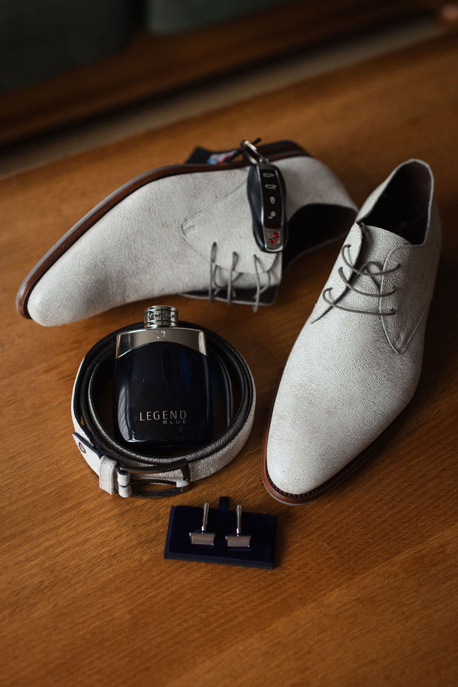 Wedding accessories including grey suede dress shoes, cologne bottle and cufflinks arranged on wooden surface.