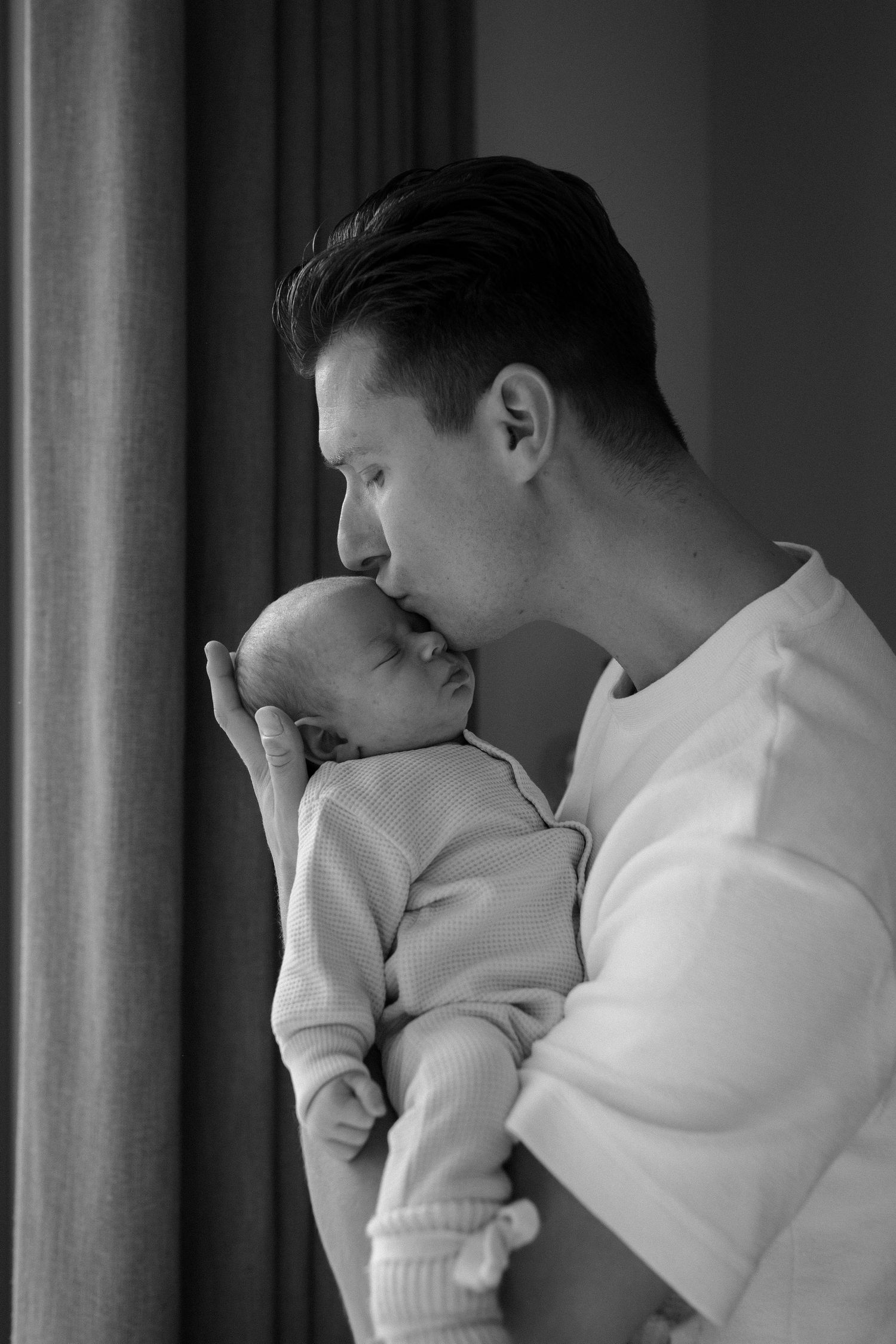 Black and white intimate portrait of parent and sleeping newborn by window.