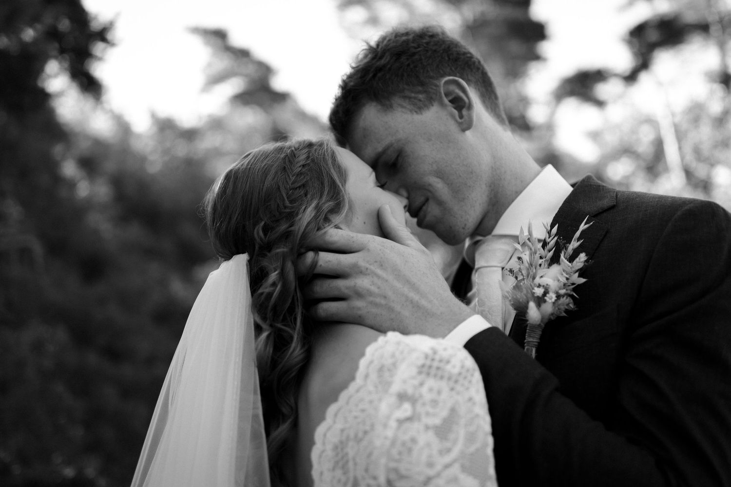 A romantic black and white wedding moment captured outdoors with a veiled bride and groom sharing an intimate kiss.
