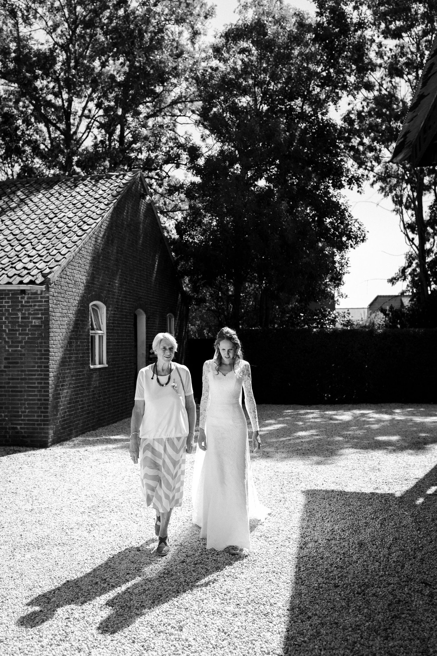 Black and white photo of two people walking together on a gravel path next to a brick building in dappled sunlight.