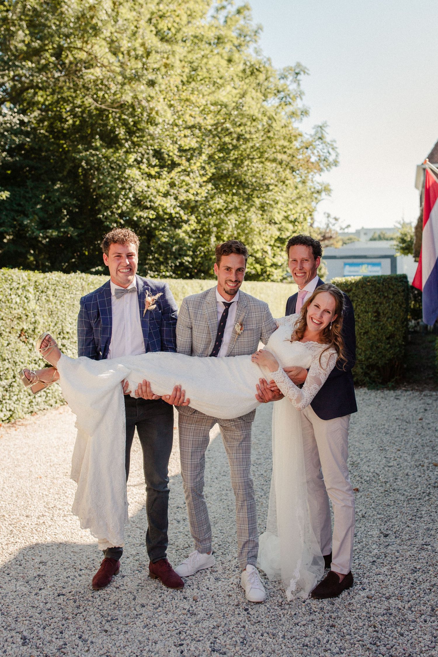 Wedding party poses playfully outdoors on a gravel path with trees and hedges in the background on a sunny day.