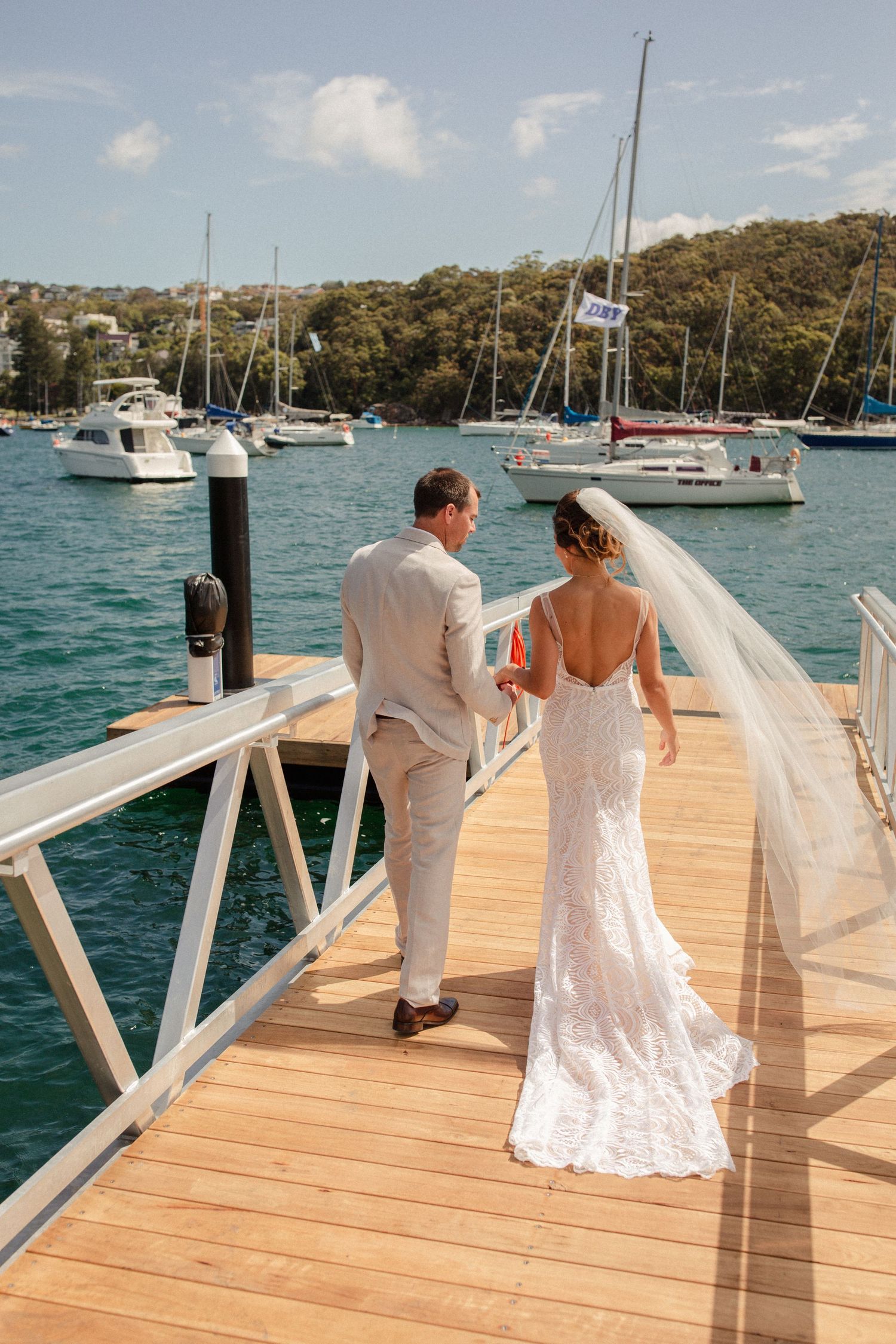 A bride and groom walk along a wooden dock with boats and blue water in the background.
