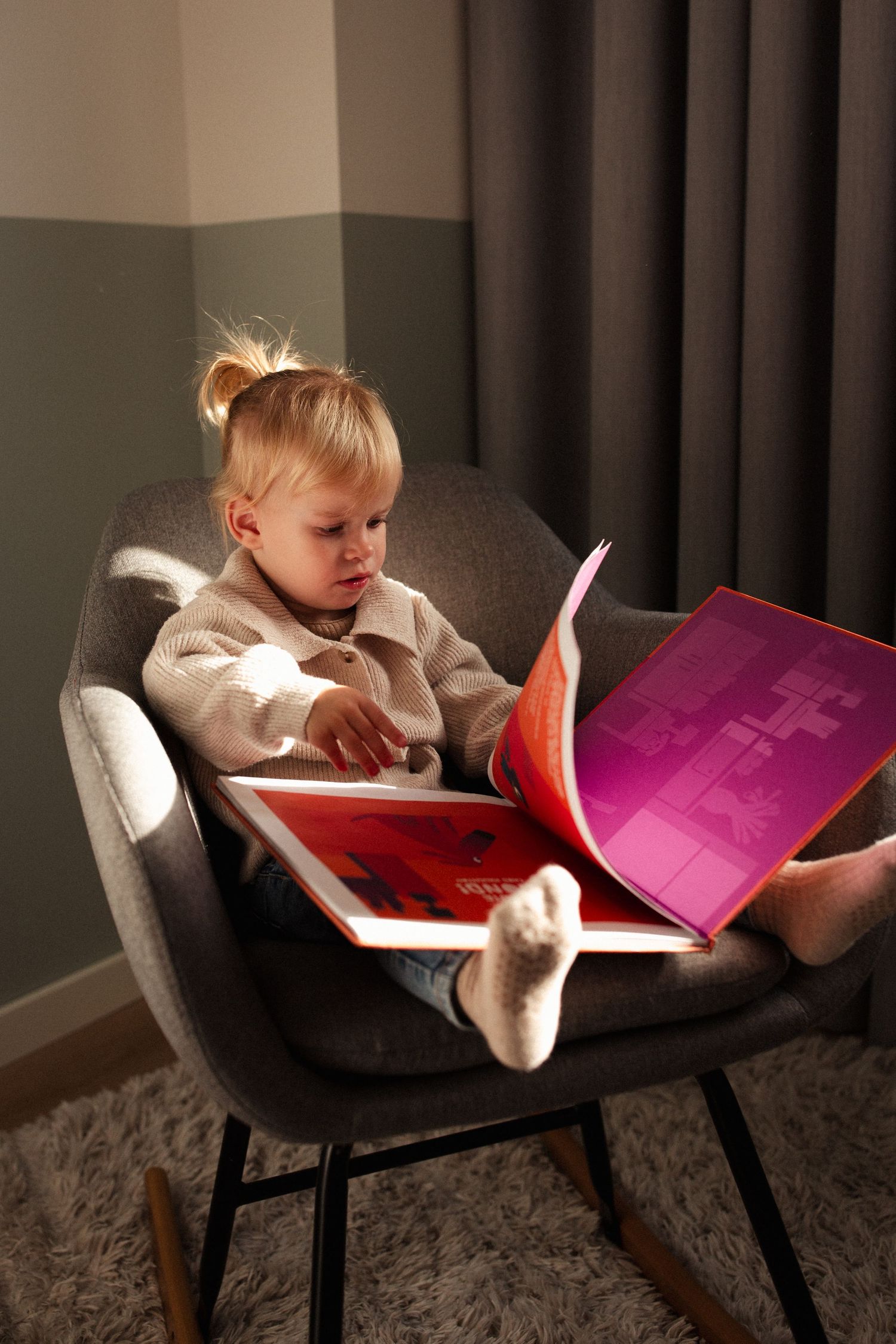Child reading colorful book while sitting in grey rocking chair.