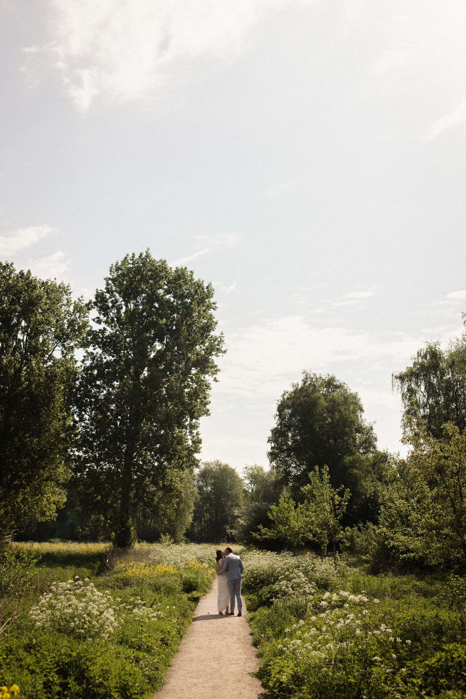 A couple walks down a winding path through a lush summer meadow surrounded by tall trees in natural light.