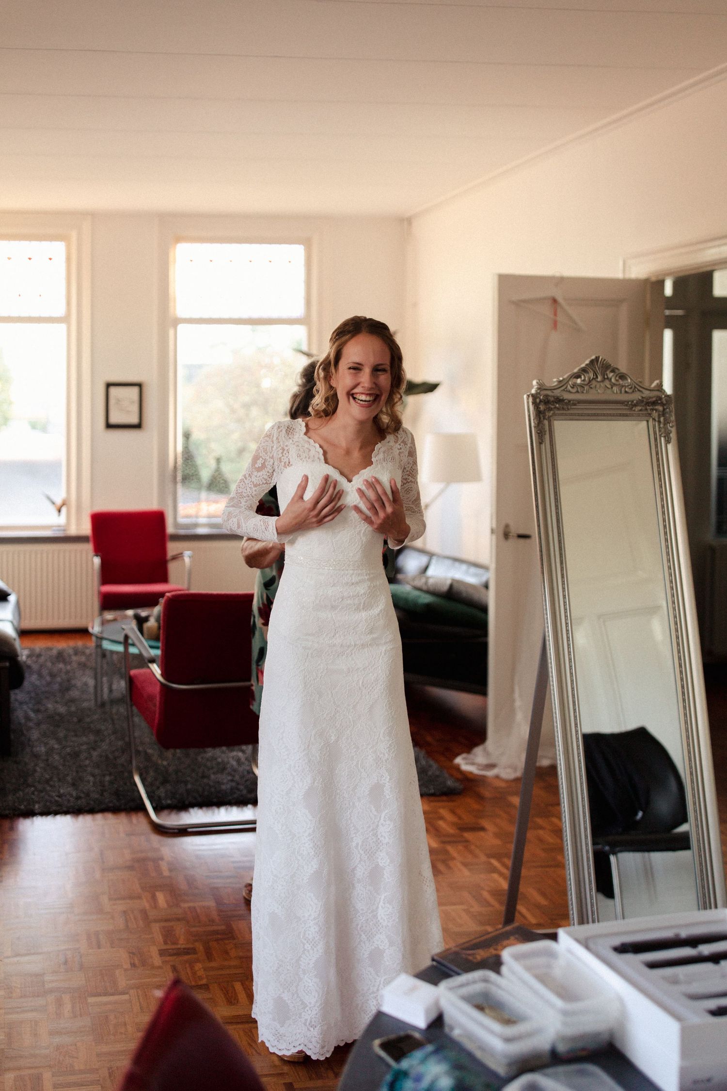 A woman in an elegant white lace wedding dress stands smiling next to a full-length mirror in a brightly lit room.