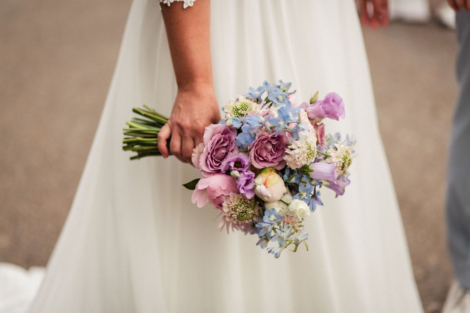 Close-up of bride holding pastel bouquet with purple roses, blue hydrangeas and pink flowers.
