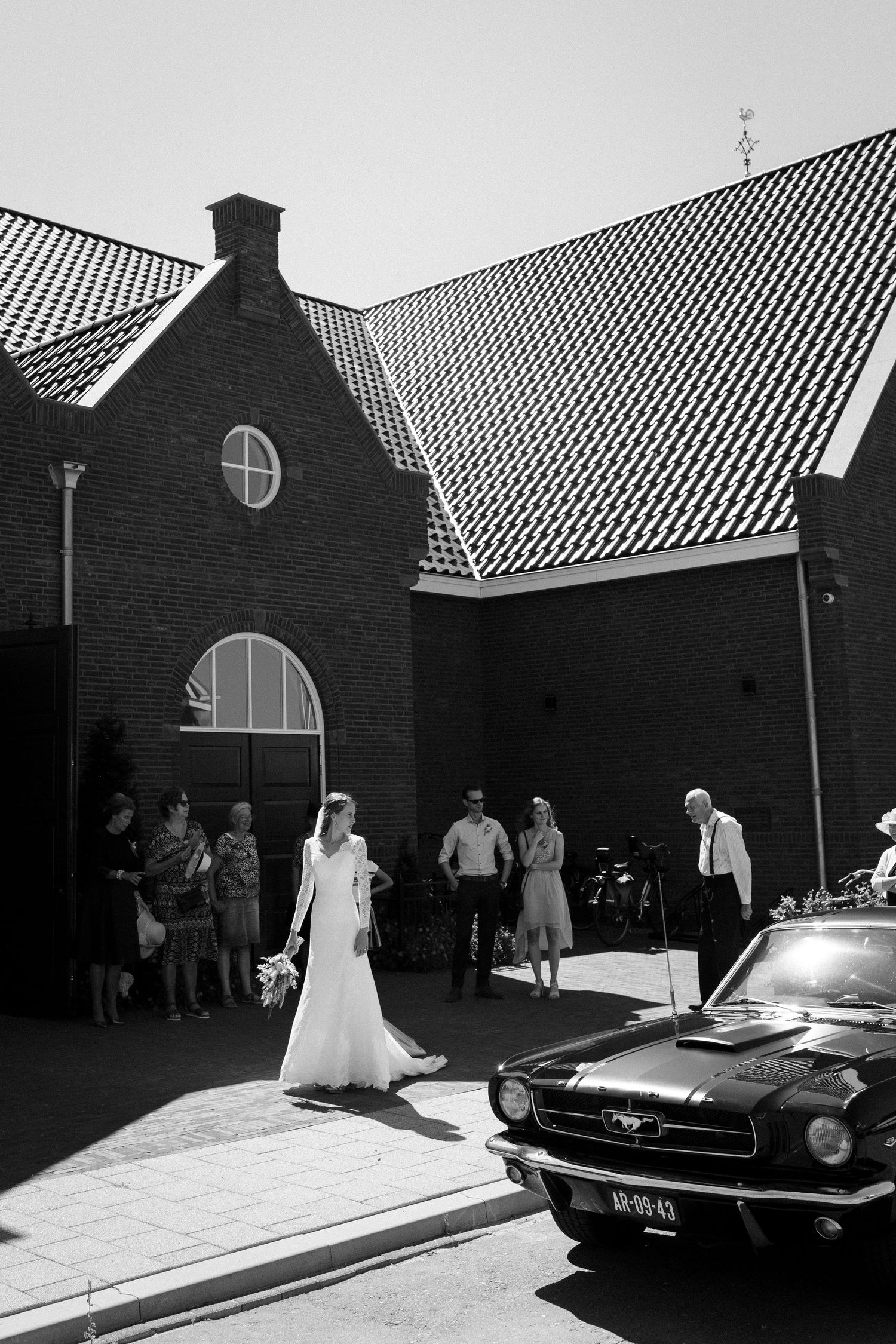 A vintage Mustang car parked outside a brick church with a Gothic-style arched entrance during a wedding ceremony.