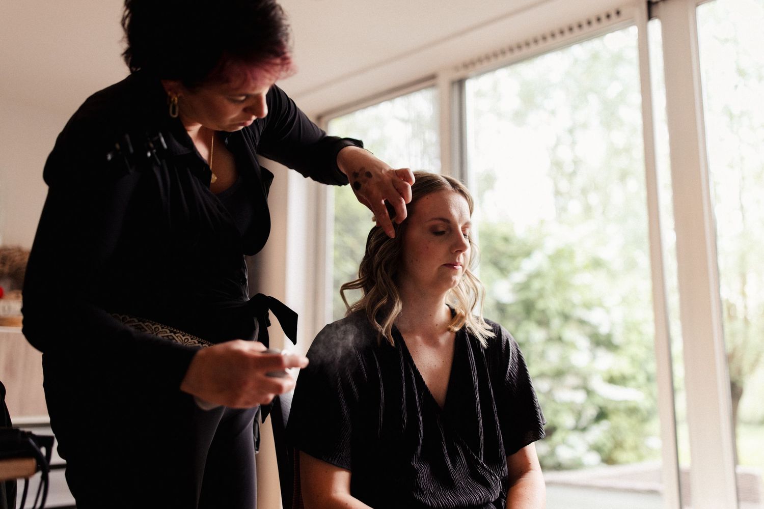 Hair stylist using tools and products while styling a client's hair in a bright, window-lit salon setting.