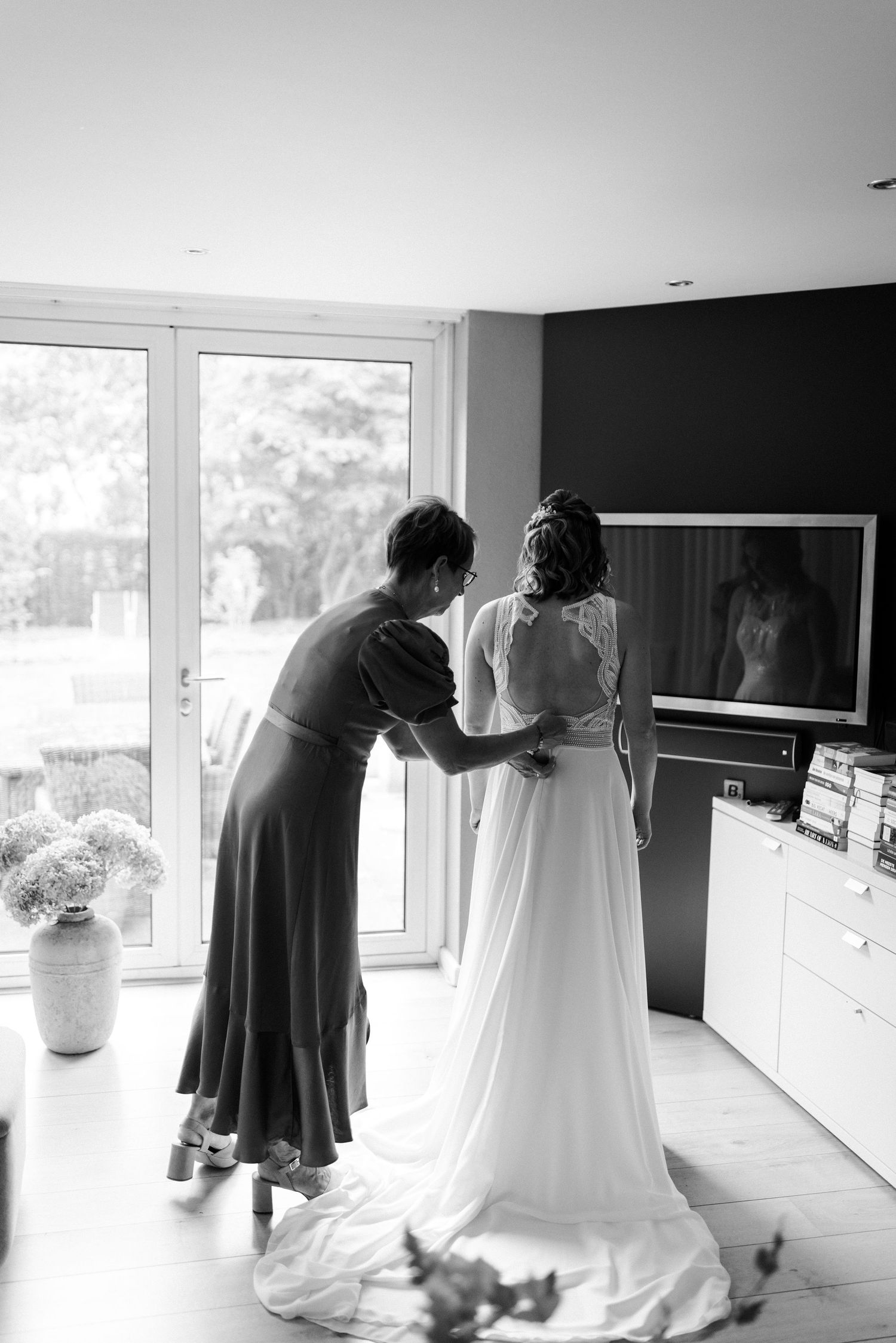 Black and white photo of a wedding dress being fitted in a bright modern room with large windows and a vase of flowers.