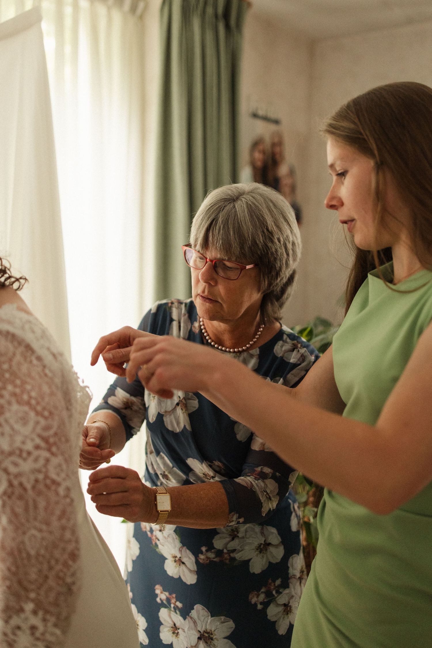Two people helping with final adjustments of wedding day preparations in a sunlit room.