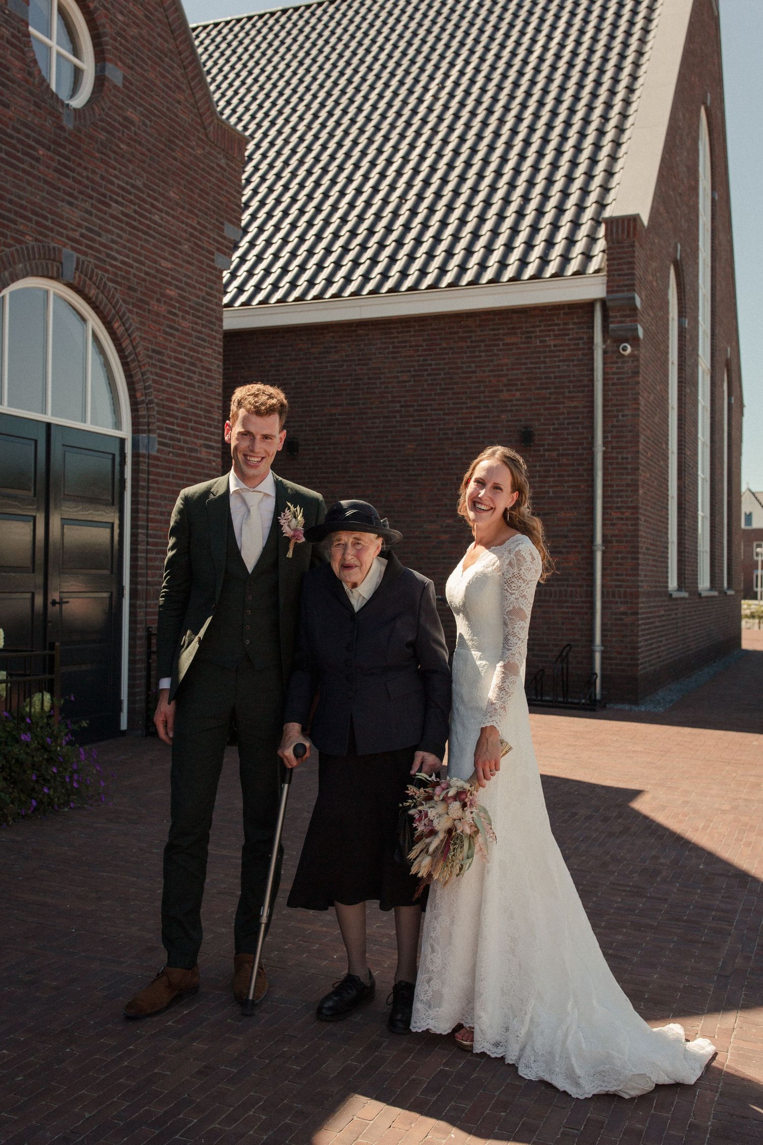 Three people stand outside a brick church building with decorative tiled roof on a sunny wedding day.