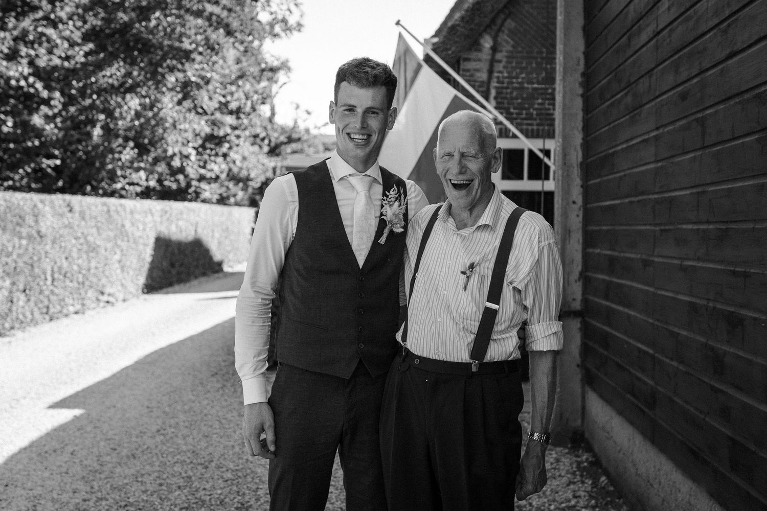 A vintage black and white photo shows two people in formal attire standing beside a building on a sunny day.