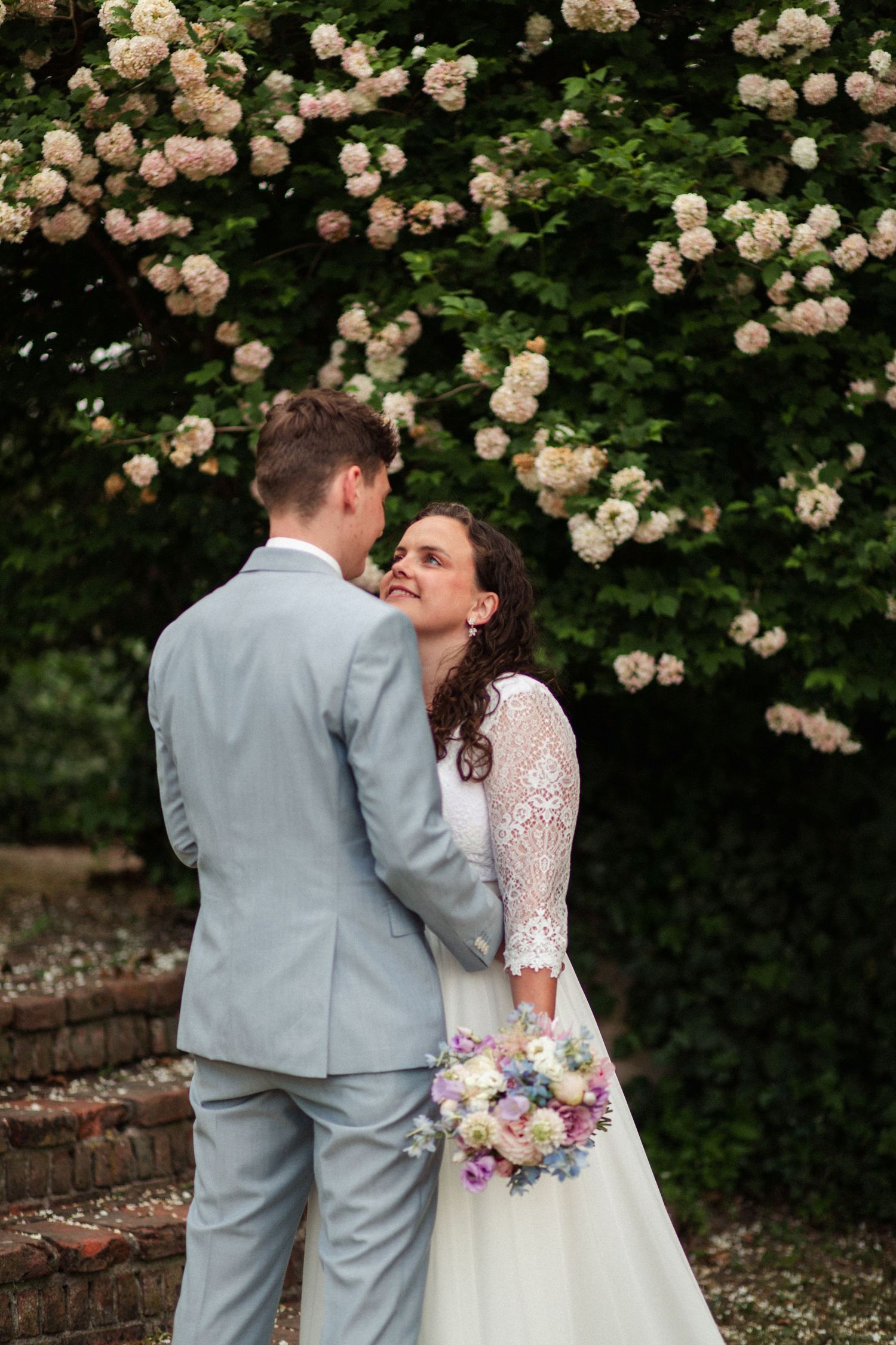A romantic wedding photo with roses blooming in the background and a pastel bouquet.