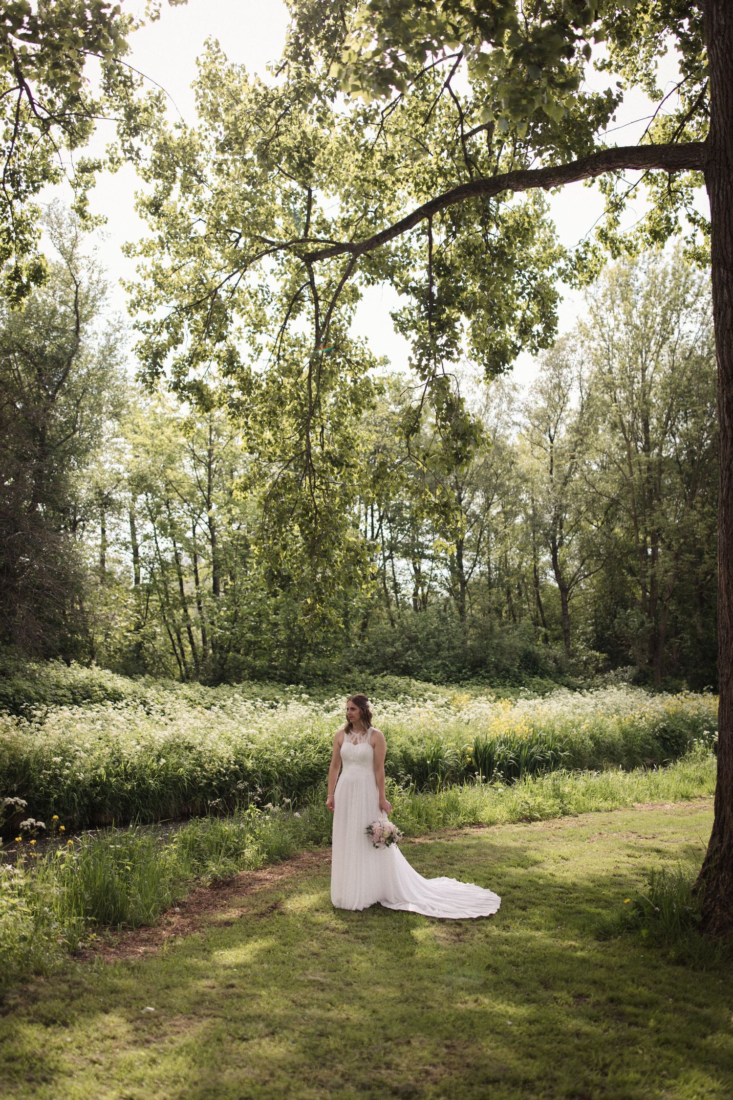A person in a flowing white wedding dress stands on a grassy path surrounded by lush summer greenery and trees.