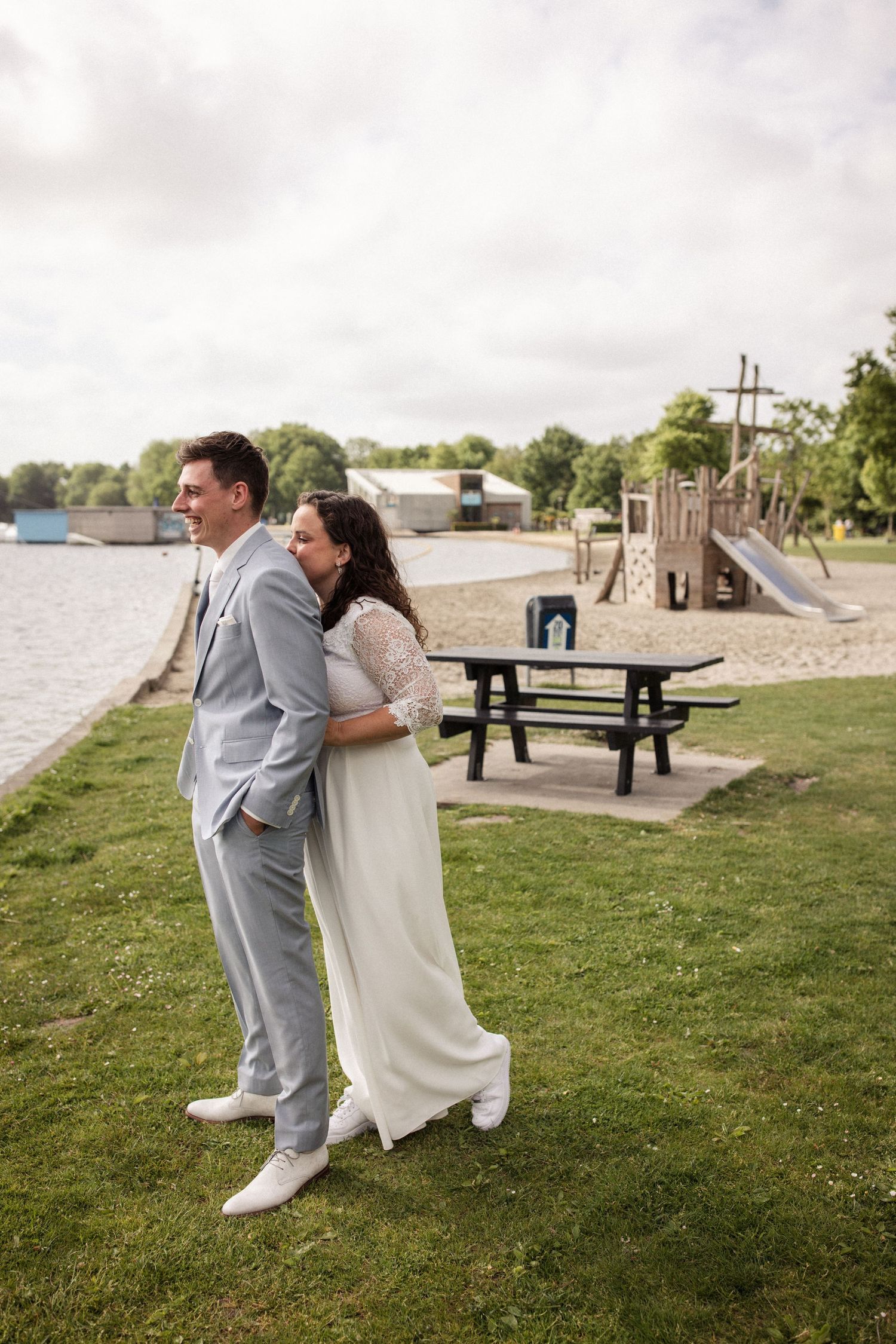 Wedding couple in formal attire embracing by a lakeside park with playground in background.