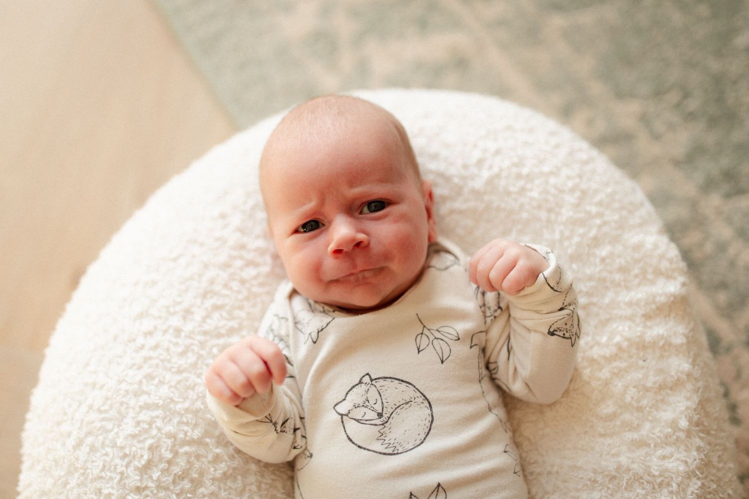 Baby wearing a onesie with animal prints lies on a round white cushion.