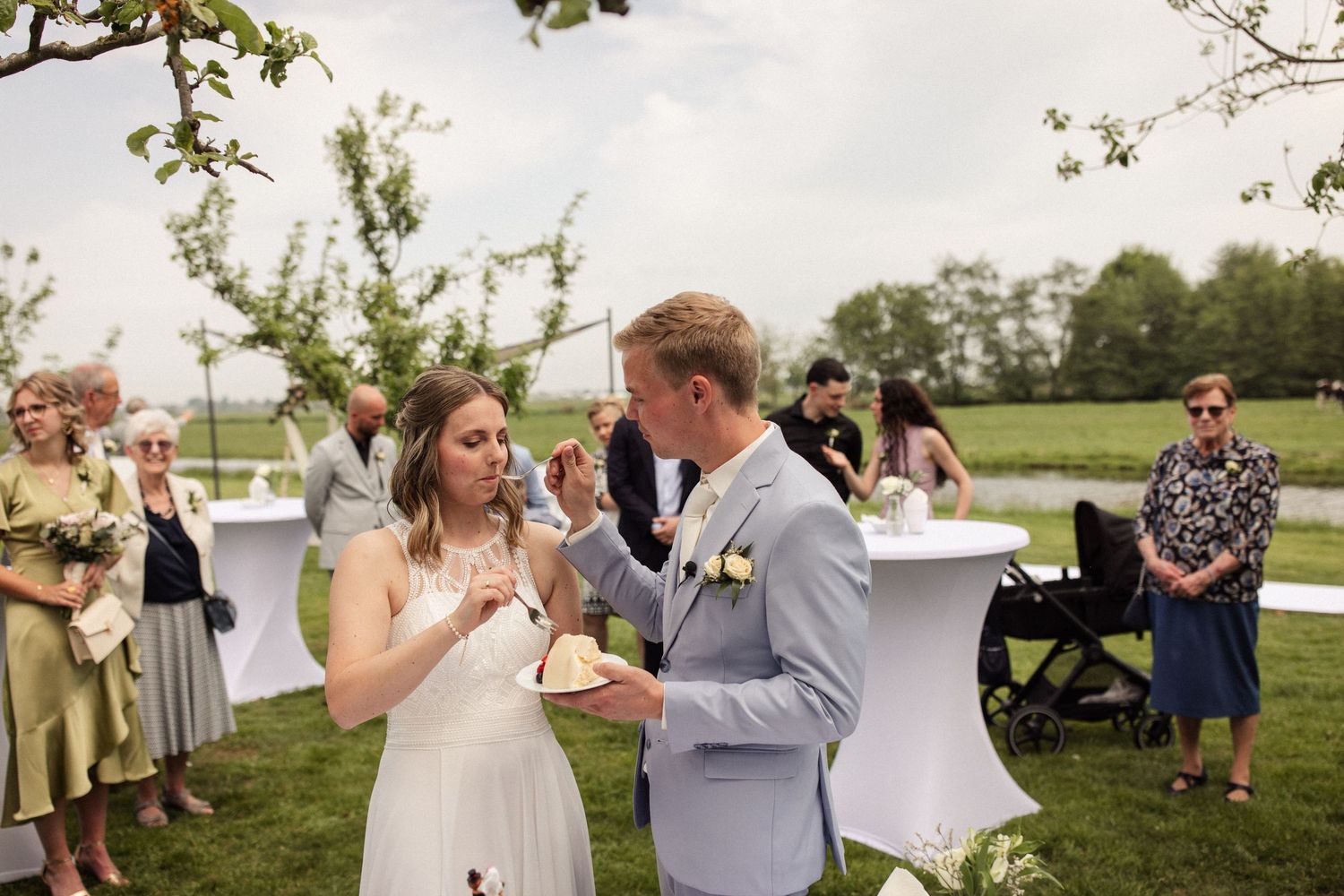 Two people share a slice of wedding cake during an outdoor garden reception with guests looking on in the background.