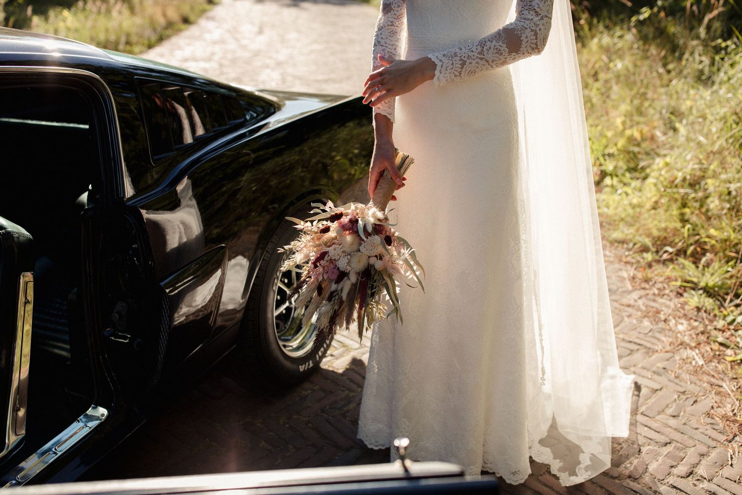 A vintage black car and flowing white wedding dress are captured in warm sunlight on a rustic path.