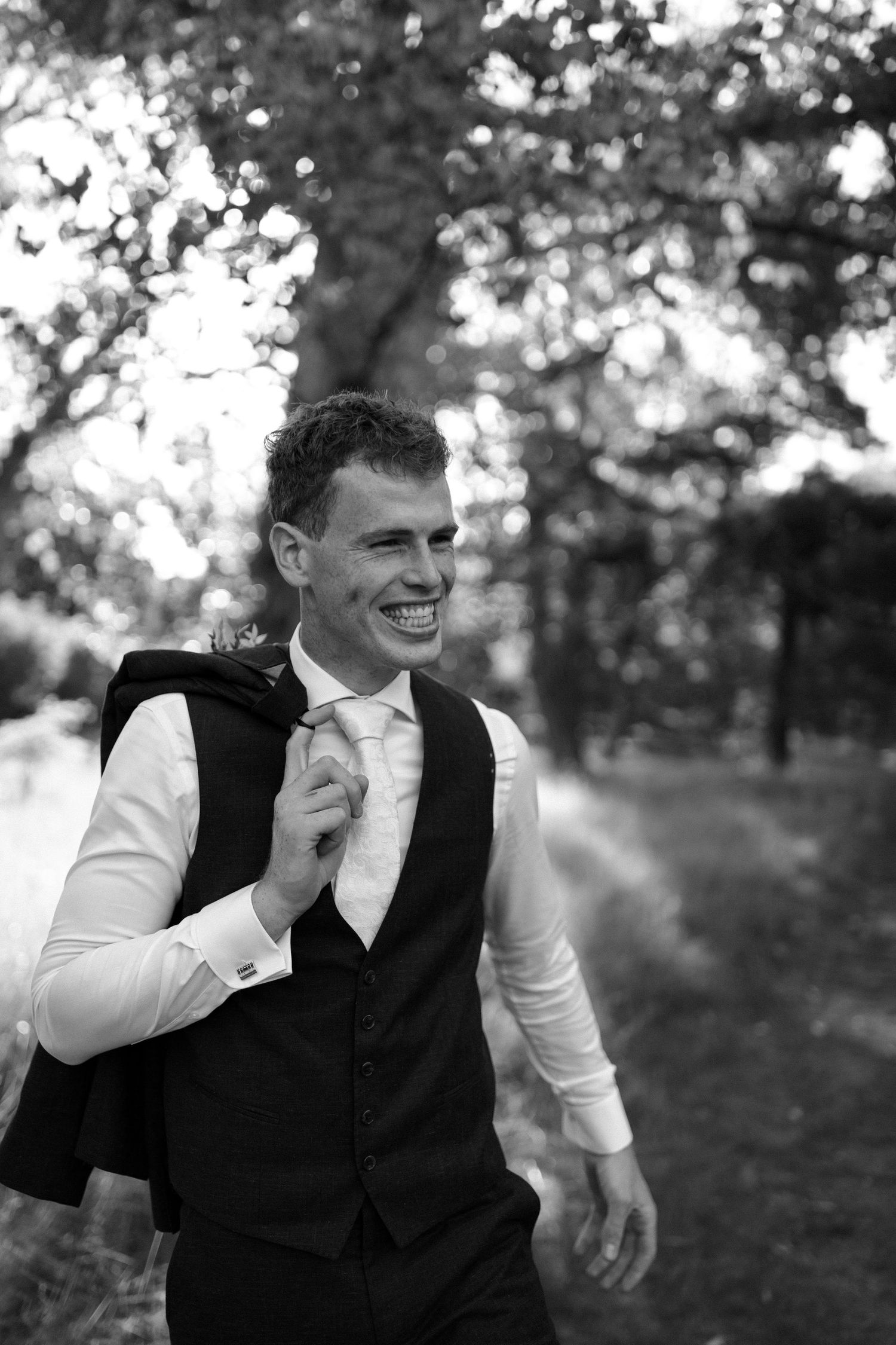 Black and white photo of a joyful person wearing a vest and tie in an outdoor setting with trees in background.