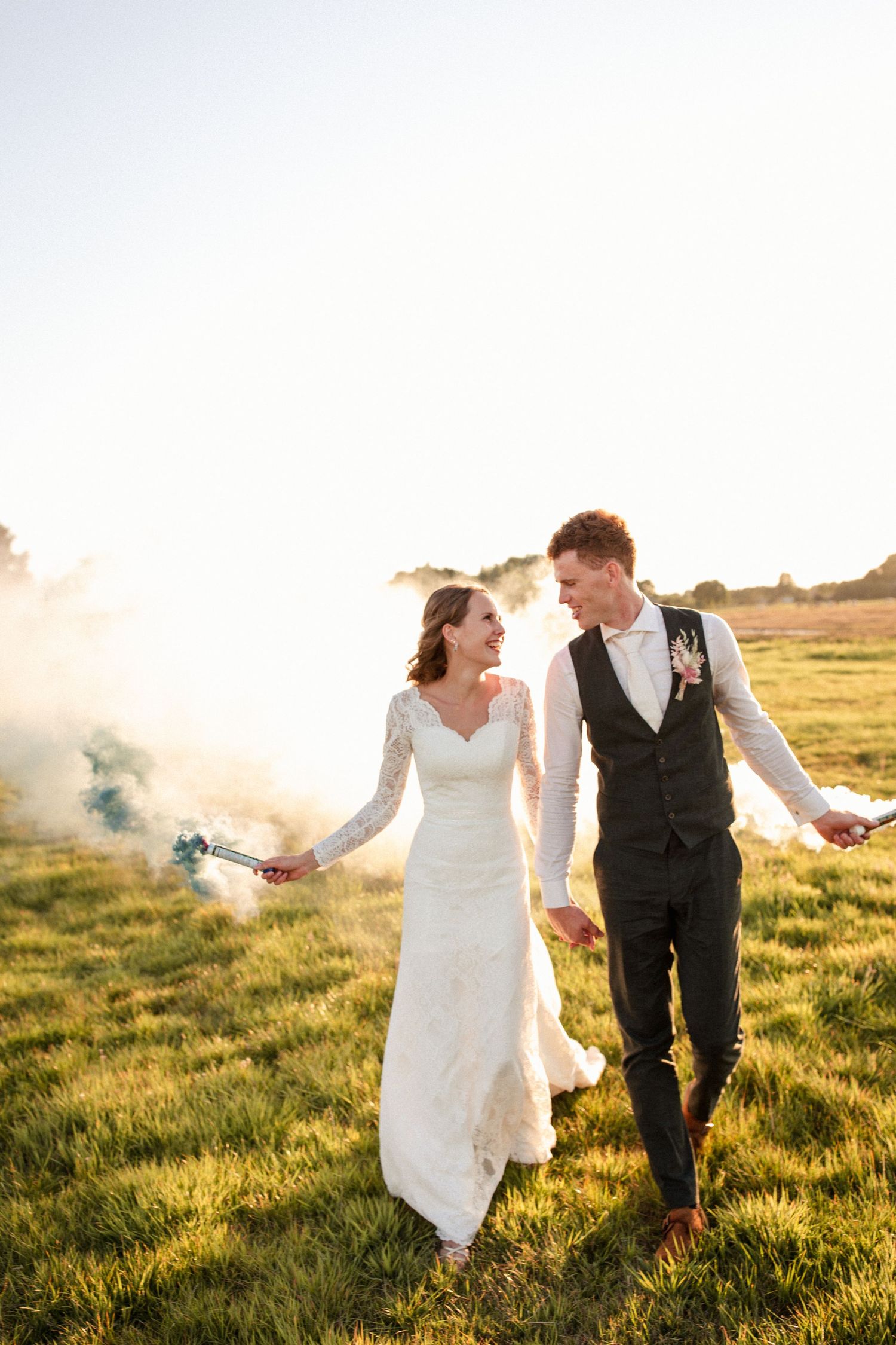 Wedding couple holding smoke flares while walking through a sunlit meadow at golden hour.